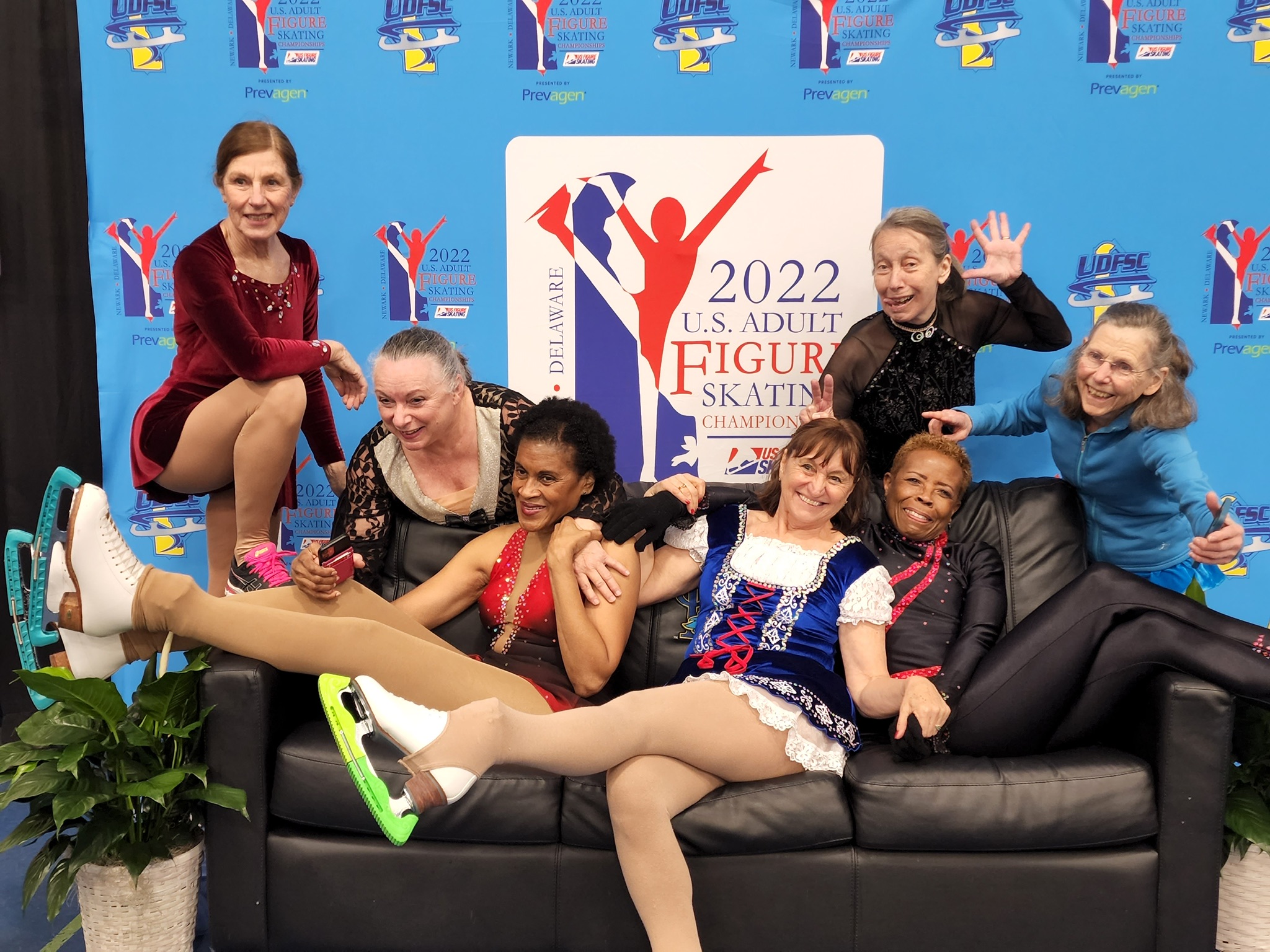 A group adult skaters sitting in their costumes on a couch in front of the 2022 Adult Championships logo making funny faces.