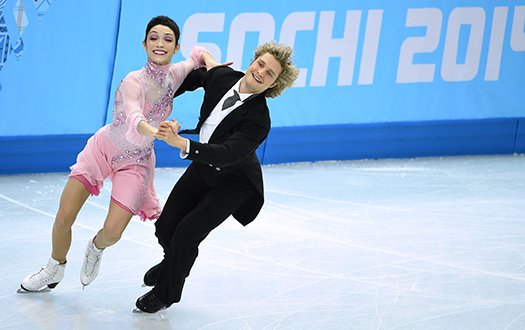 Meryl Davis, wearing pink, and Charlie White, wearing a suit, skate in front of the Sochi 2014 banner, while performing a step sequence. 