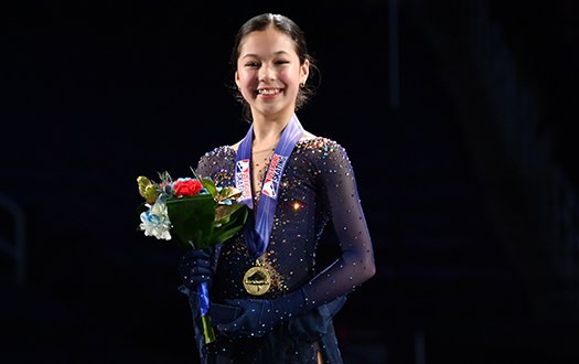 Alysa Liu with her medal atop the podium, smiles for the camera. 