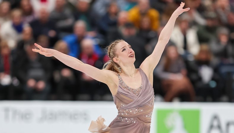 Amber Glenn looks upward with her arms outstretched. She is a young woman wearing her hair back in braids. She is wearing a light pink skating costume