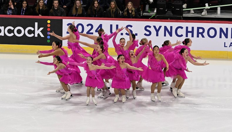 Teams Elite juvenile performs wearing matching bright pink costumes. They all stand in a circle, facing outward with their arms outstretched and excited looks on their faces. 