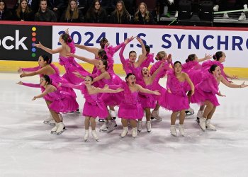 Teams Elite juvenile performs wearing matching bright pink costumes. They all stand in a circle, facing outward with their arms outstretched and excited looks on their faces. 