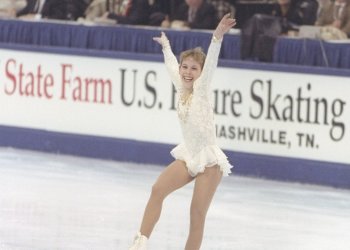 Tara Lipinski in a white lacy dress with her arms above on the ice in front of the 1997 State Farm U.S. Figure Skating Championships rinkboard.