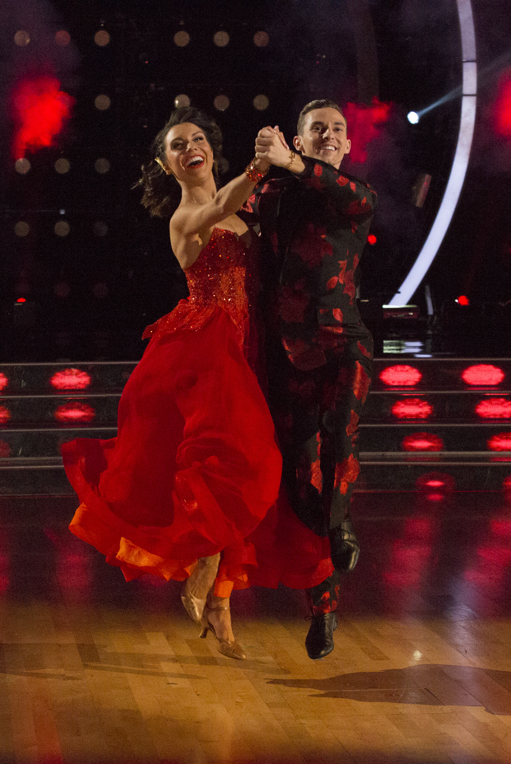 Adam Rippon dances with his female partner on Dancing with the Stars. His partner is wearing a floor length strapless dress and Adam is wearing a red and black suit