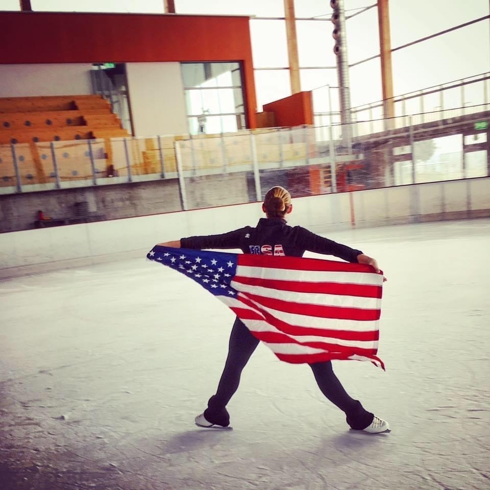 Casey Ouellette performs a spread eagle wearing a Team USA jacket with the American flag draped along her back.