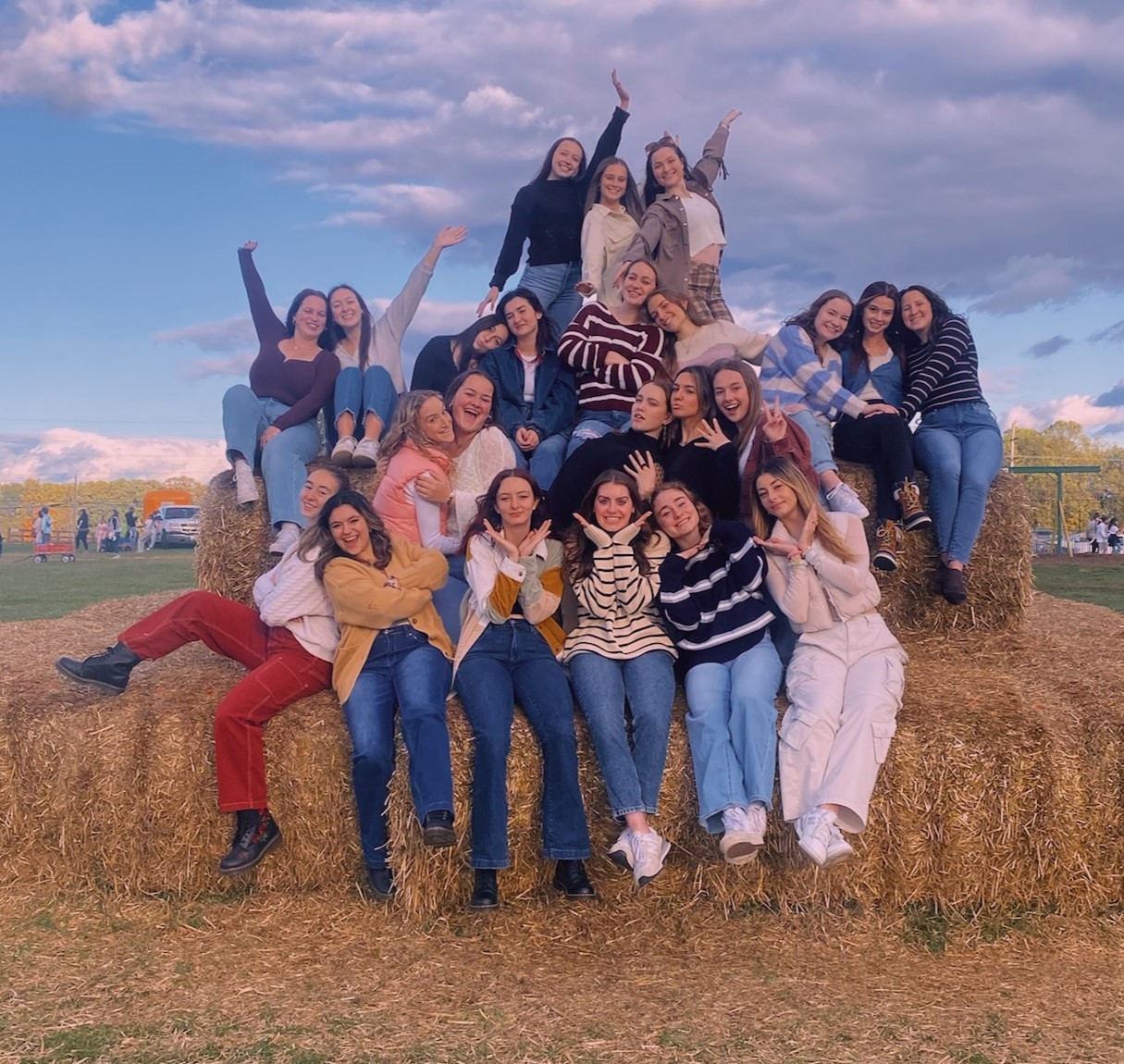 UD poses for a group photo with their hands in the air while sitting on a large bale of hay at a pumpkin patch