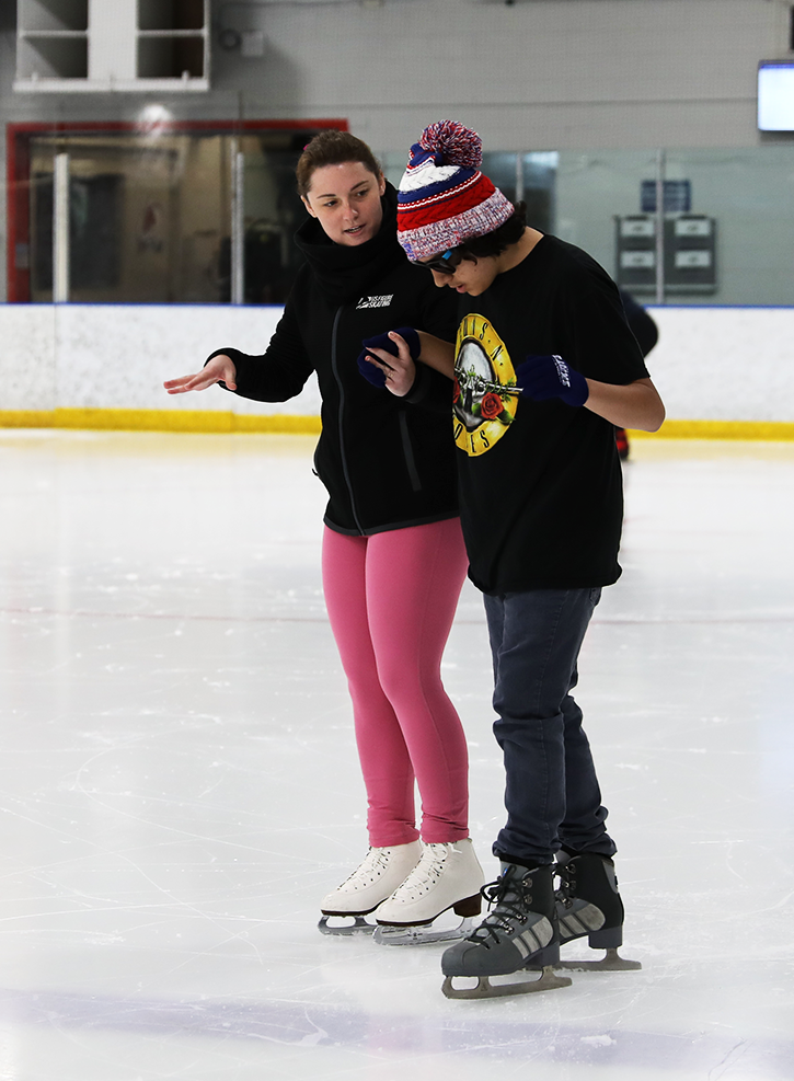 A woman stands with a student giving him pointers on how to skate while on the ice. The woman (L) is a white woman with long brown hair. She is wearing a black jacket with bright pink pants. The student is Latino with longer black hair. He is wearing a U.S. Figure Skating red white and blue knit hat, sunglasses and and a Guns N Roses tshirt