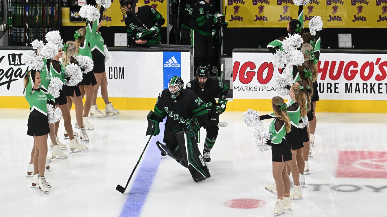 UND hockey cheerleaders welcome the UND hockey team to the ice. The cheerleaders are wearing long sleeve green, white and black uniforms and the hockey players are wearing hockey uniforms in the same colors