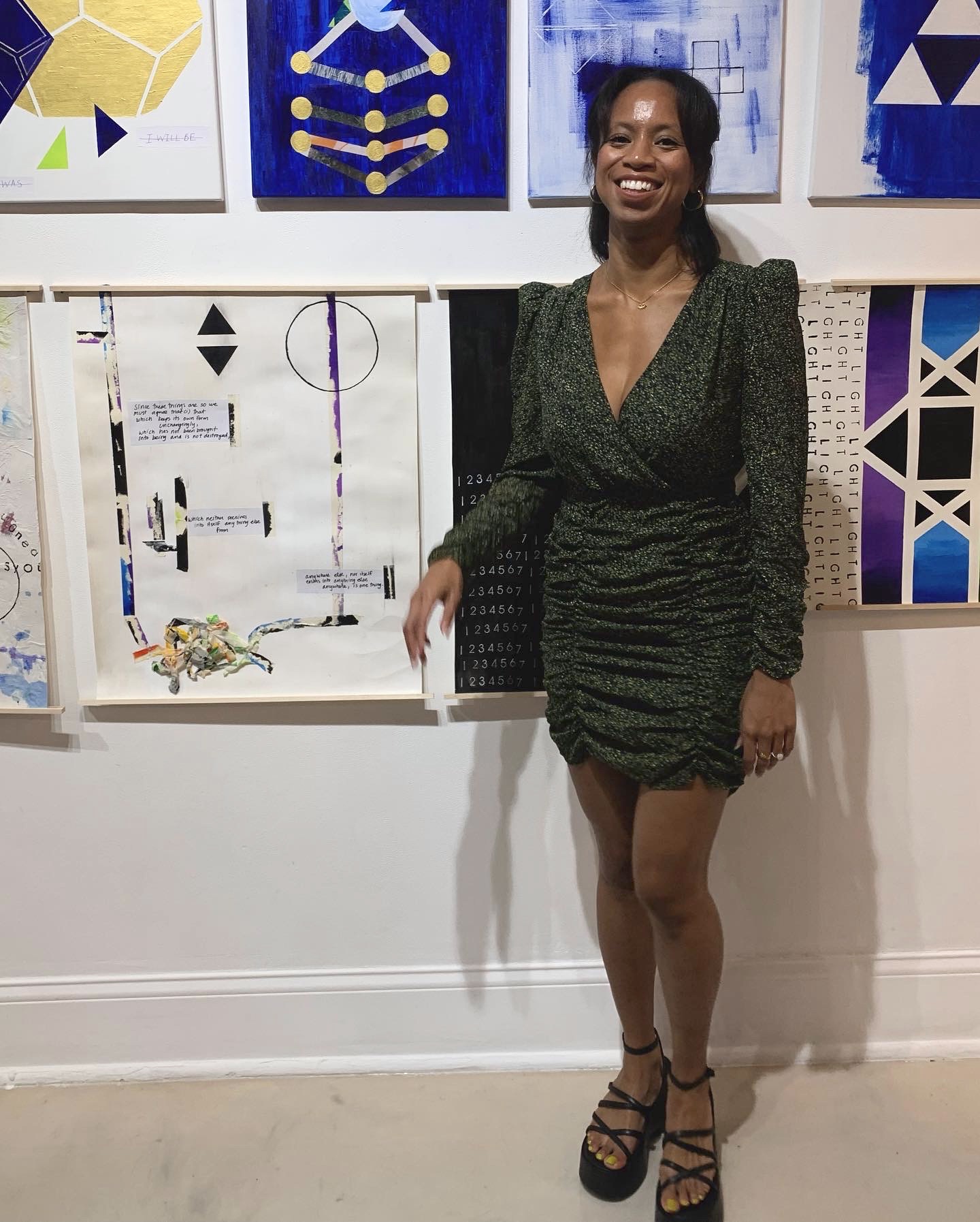 Musademba stands in front of a wall displaying some of her art. She is a Black woman wearing a dark green dress with a v-neck and strappy black sandels