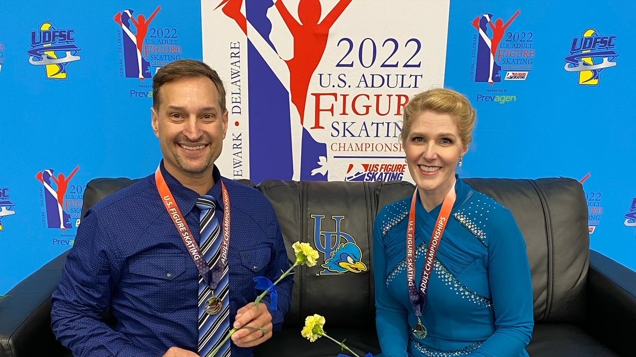 John and Janet Stone sit on a black couch in front of a large logo for the 2022 U.S. Adult Figure Skating Championships. John (left) is a middle aged white man weating a blue dress shirt and blue tie. He wears a U.S. Figure Skating Adult Championships medal arounf his neck. Janet (left) is a middle aged white woman with blonde hair tied back in a bun. She is wearing a turquoise long sleeve skating costume. She has a medal around her neck as well. Both are holding yellow flowers. 