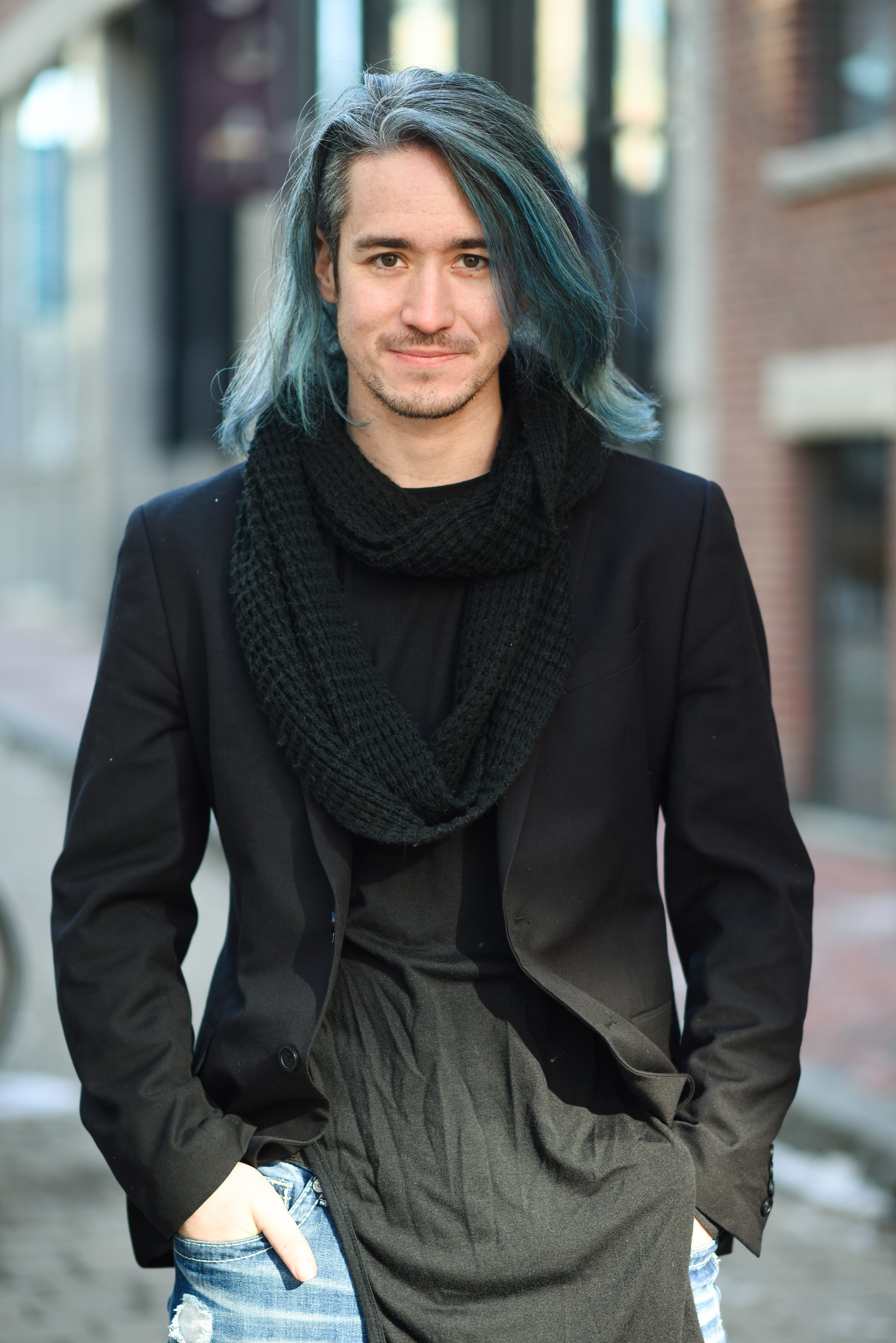 Professional photo of Adam Blake. Adam is a man with shoulder length blue hair. He is wearing a black shirt, black blazer, black scarf and blue jeans