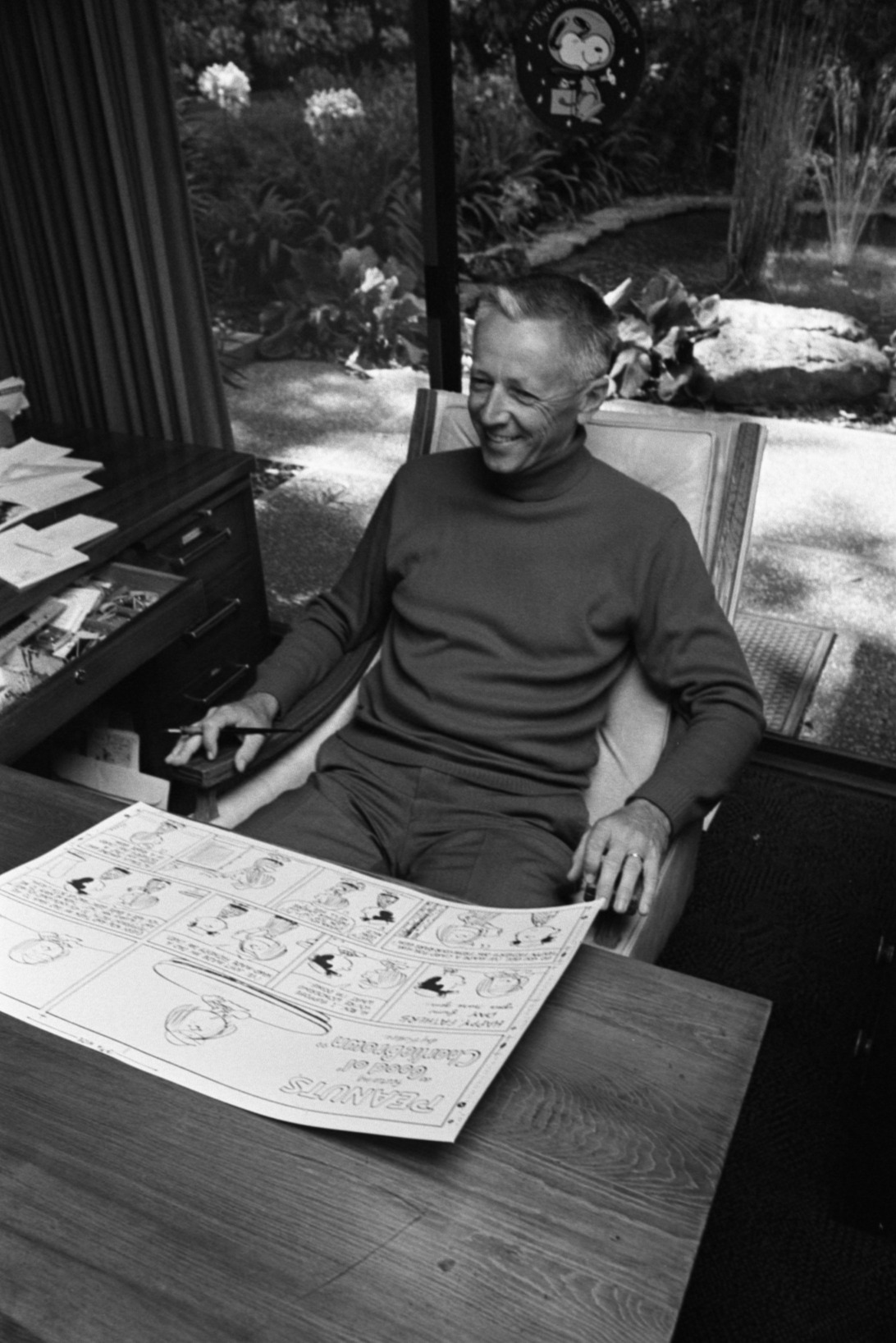 Charles Schulz sits in a chair smiling, a drawing of his Peanuts comic sitting at the table he's sitting at.