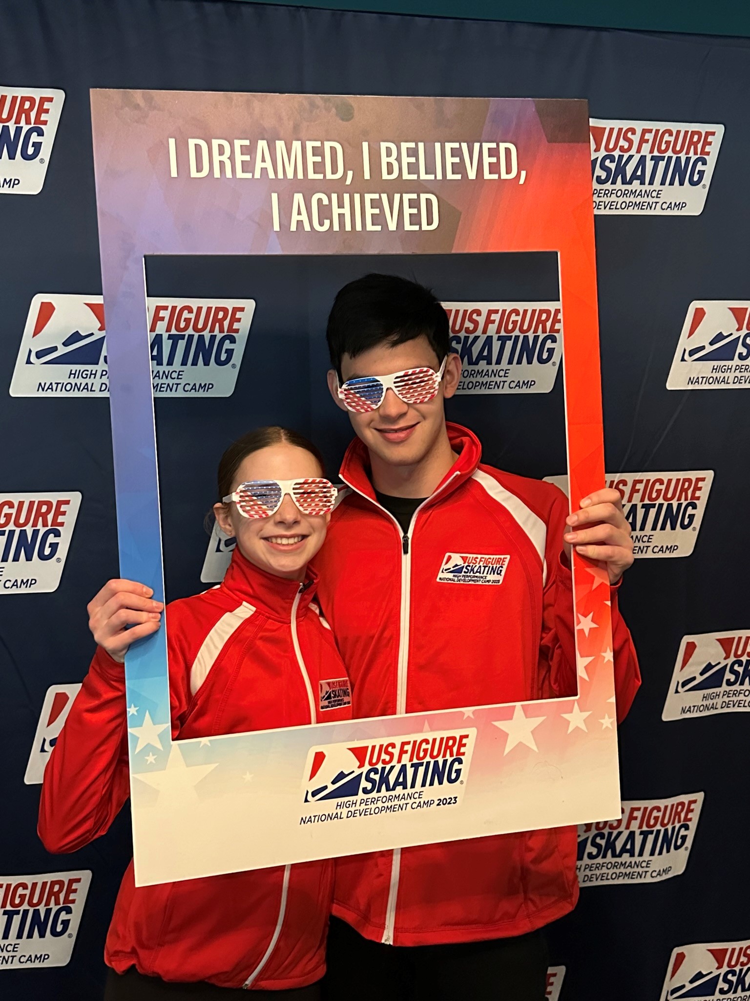 Clara Finkelstein and Mark Williams pose with a picture frame that says "I dreamed, I believed, I achieved" and funny red white and blue sunglasses  Clara is a young white girl with brown hair tied back and Mark is a tall white boy with shirt black hair. They are both wearing red National Development Team jackets