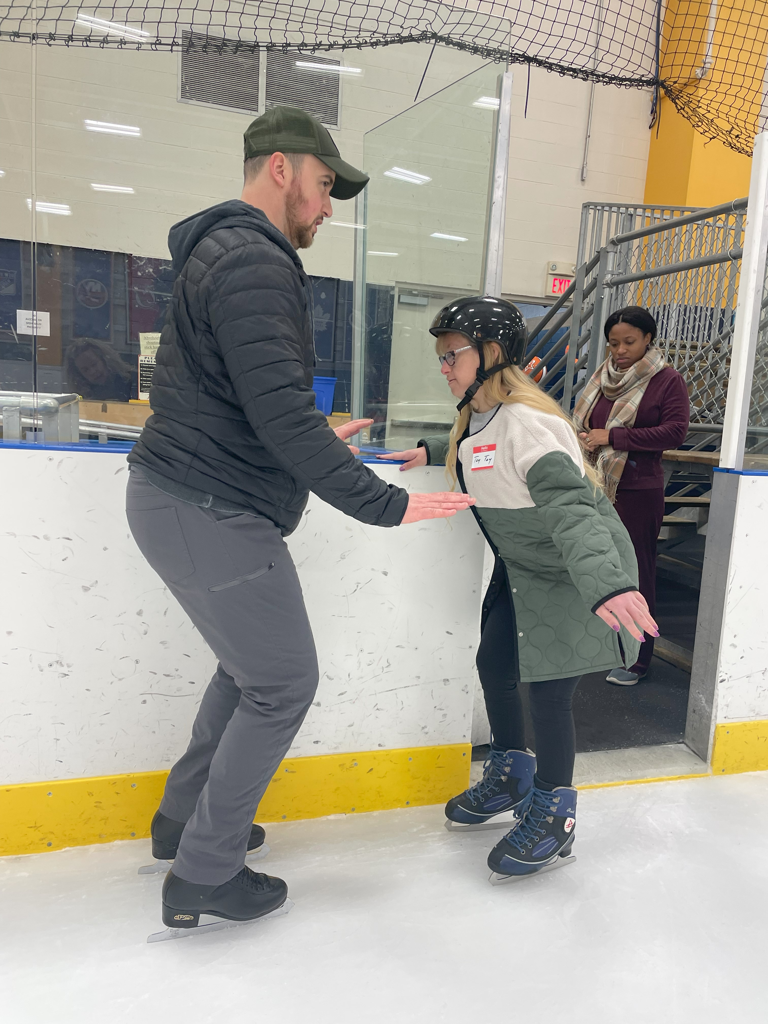 A coach helps a skater balance on the ice. The coach (left) is a white man with a beard wearing a baseball cap and black winter jacket. The skater (right) is a woman with Down Syndrome wearing a black helmet and a white and green winter jacket. 