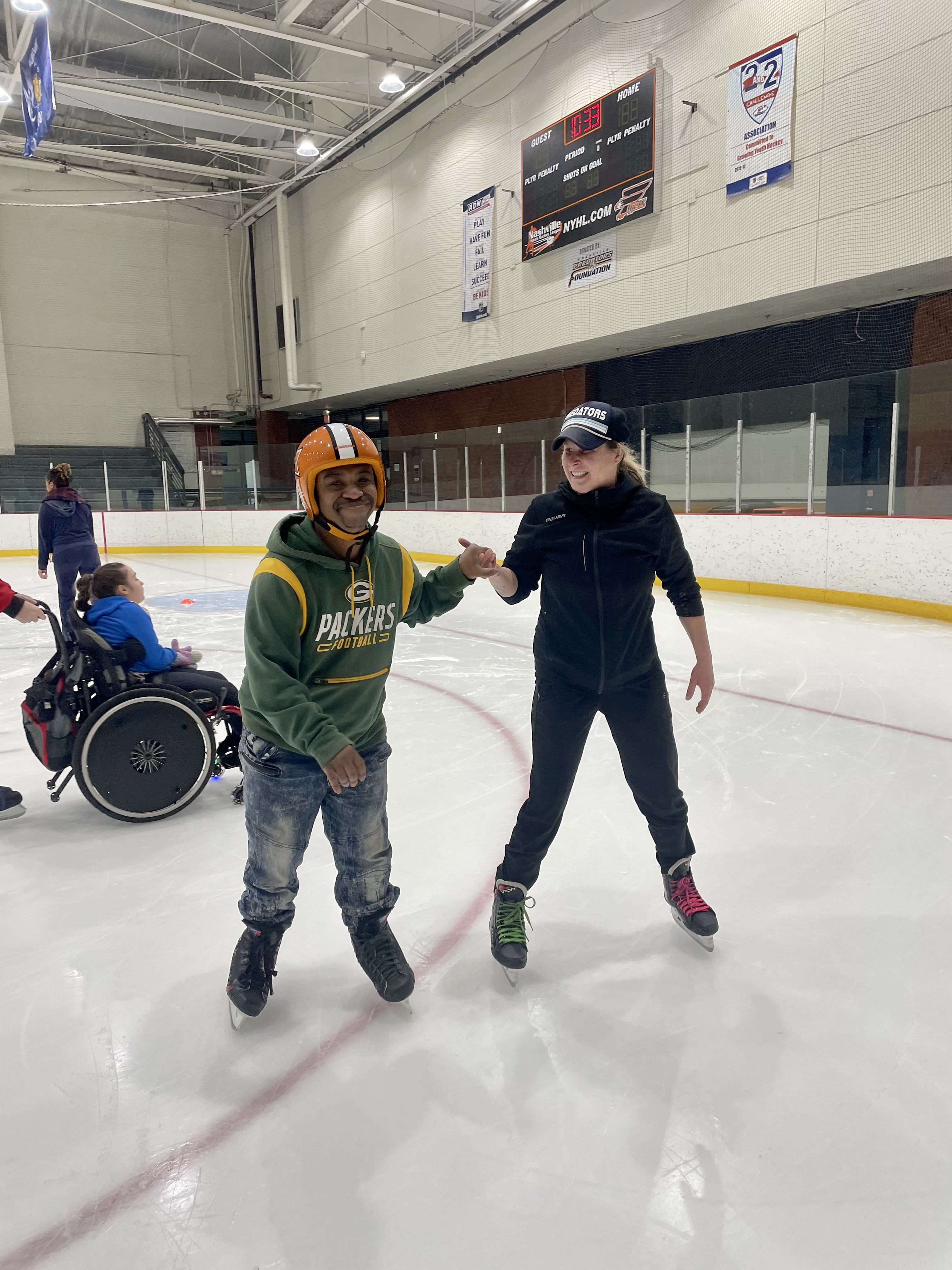 A female coach wearing a black jacket and baseball cap holds a skaters hand to help him balance on the ice. The skater is a young black man wearing a Packers hoodie, jeans and an orange helmet. A young girl in a wheelchair can be seen on the ice in the background