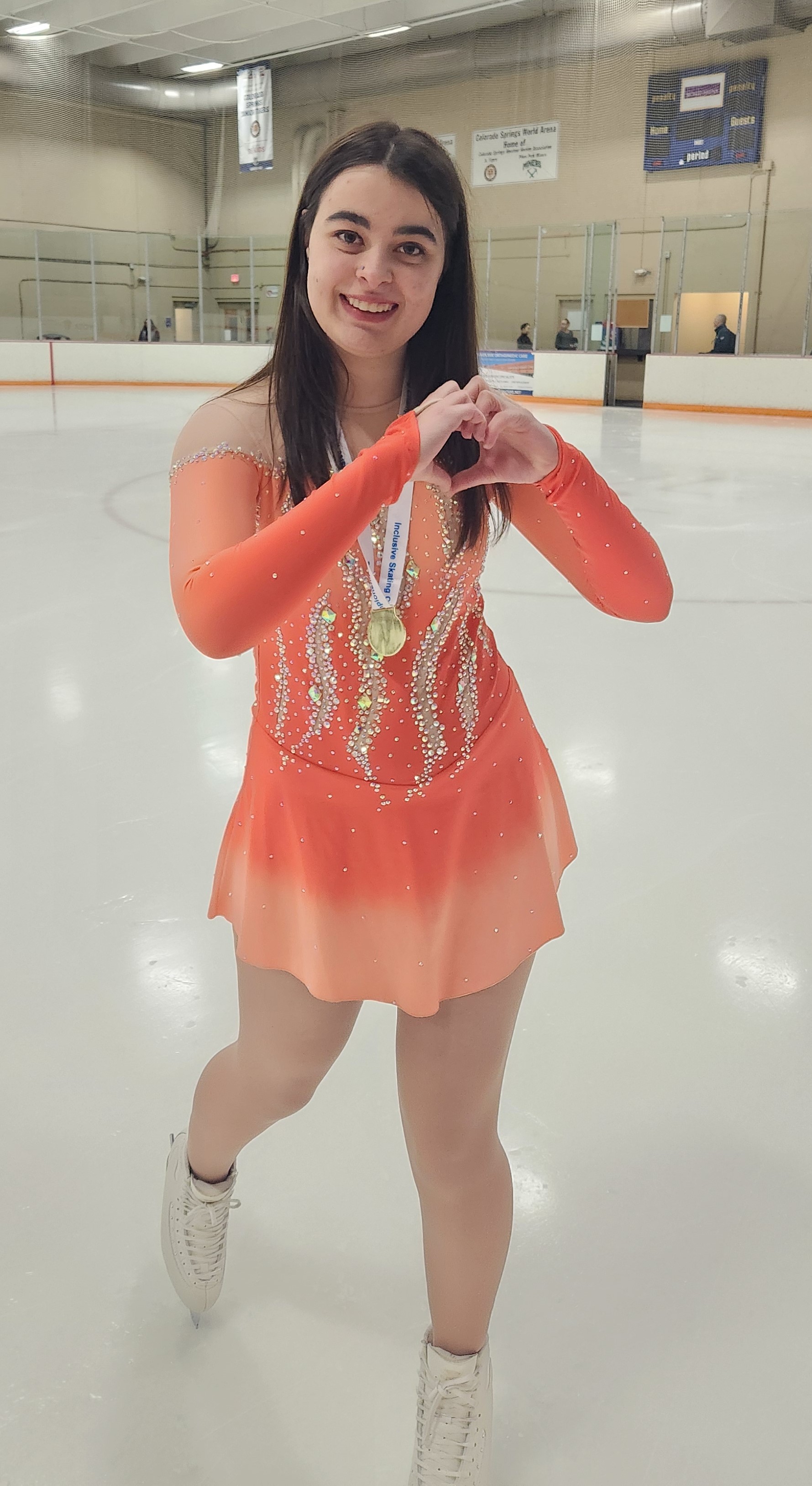 Dacia Loconte makes a heart with her hands and smiles at the camera wearing a long sleeve orange skating costume