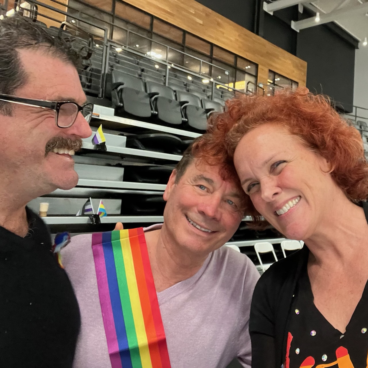 (L-R) Doug Webster, Randy Gardner, and Elin Schran pose for a selfie. Doug is an older white man with a mustache wearing glasses, Randy is an older white man wearing a pride flag across his torso, and Elin is an older white woman with red hair wearing a shirt with the Pride flag on it 