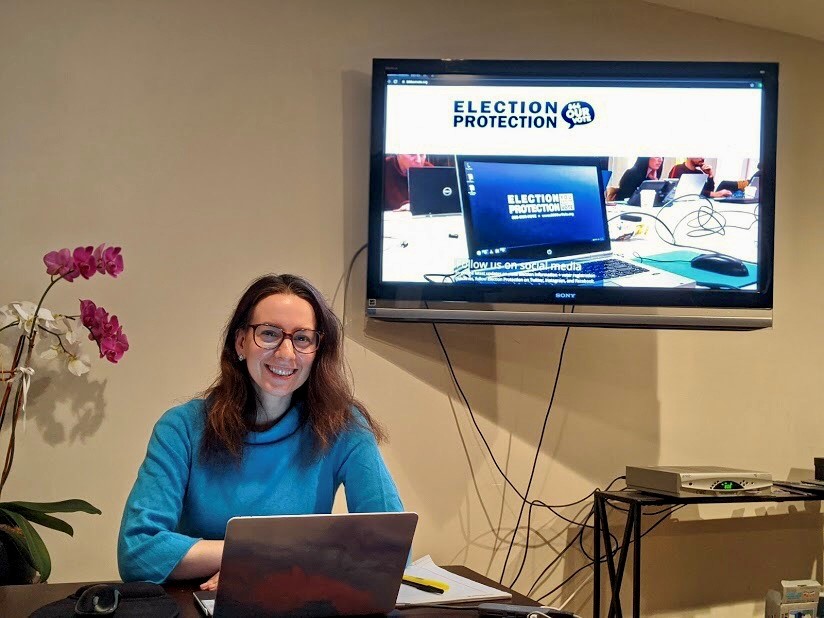 Sarah Hughes sits behind a desk with a laptop wearing glasses and a bright blue sweater.