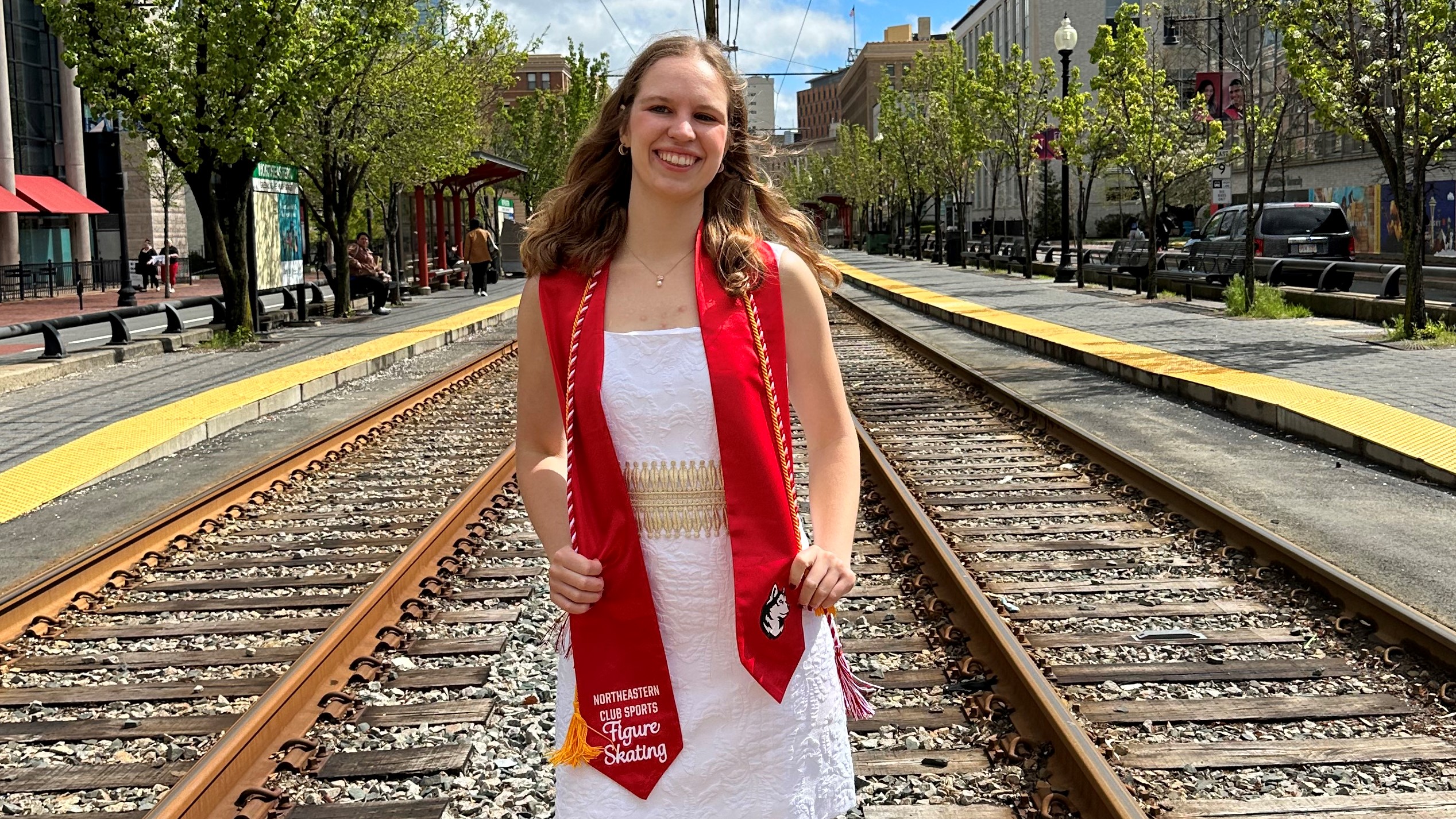 Elizabeth Klemm poses for a photo on train tracks. She is a young white woman with shoulder length blonde hair. She is wearing a white dress with a red grauation stole and a red and white graduation cord 