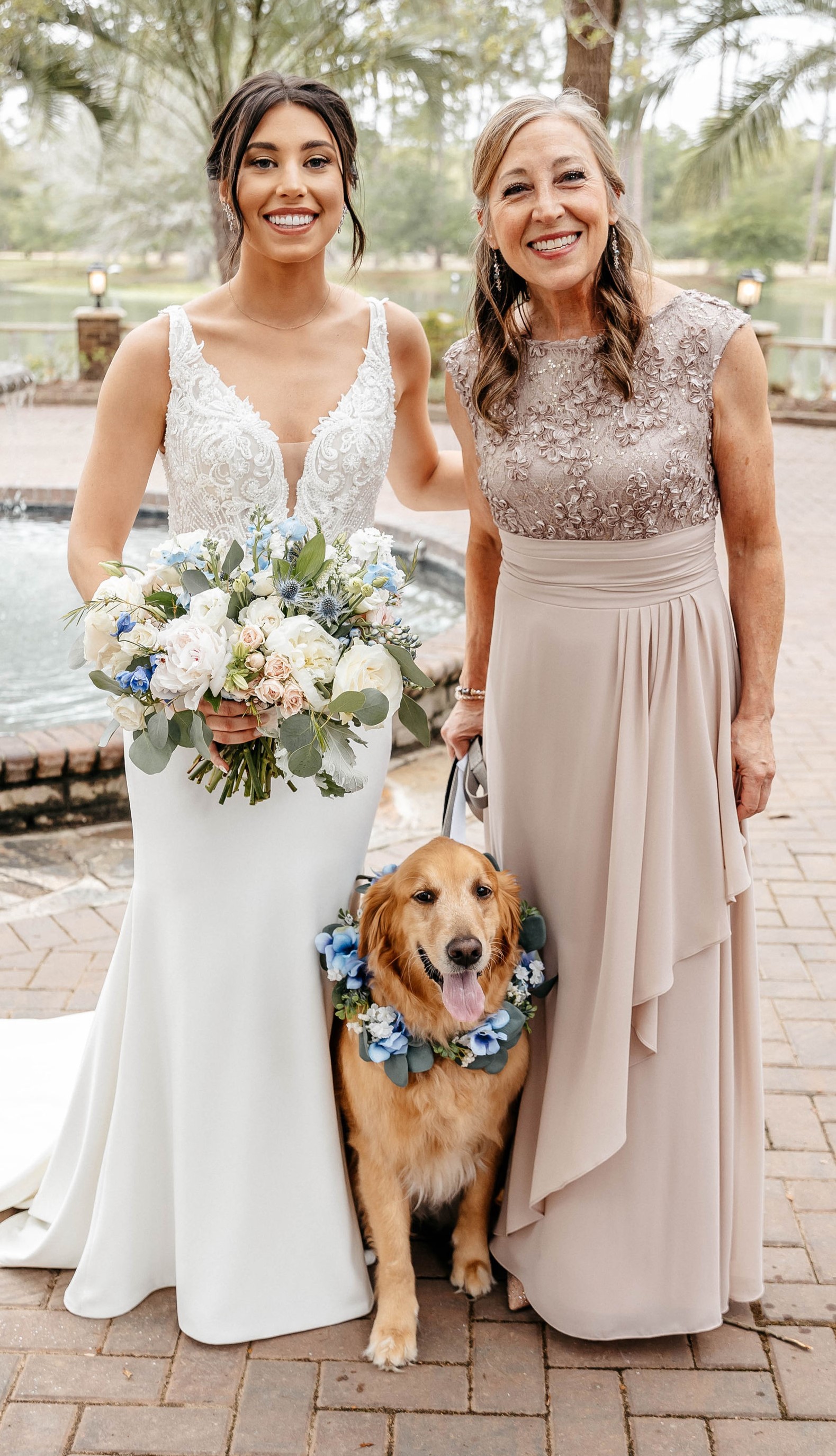 Lenz and her daughter Emma at Emma's wedding. Emma (left) is a white woman eith brown hair tied back wearing a sleeveless wedding dress and holding a bouquet of flowers. To her left is her mom Sandy wearing a tan colored and floor length dress with jewels on the top. Between them is a golden retriever with its tongue sticking out and flowers around its neck