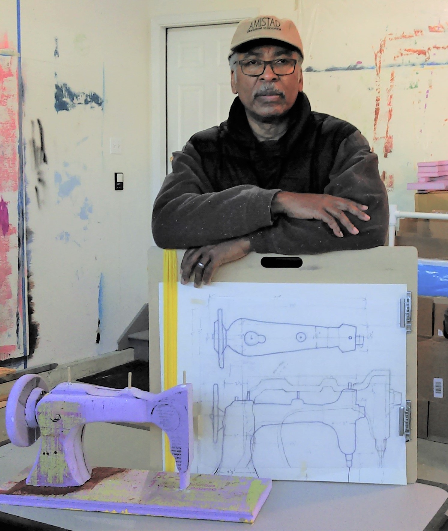 Gerald Thomas poses with a drawing he created of a sewing machine. Gerald is an older African American man wearing a black hoodie and tan baseball cap. 