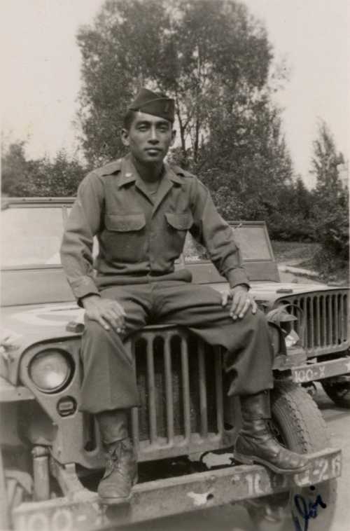 black and white photo of George Doi. He is a young Asian man wearing a military uniform. He is sitting on top of a Jeep