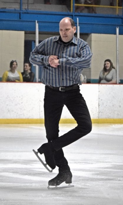 Mark Getman competes wearing a blue button down shirt with vertical white stripes and black pants. He has his arms tucked close to his body as he finishes a spin