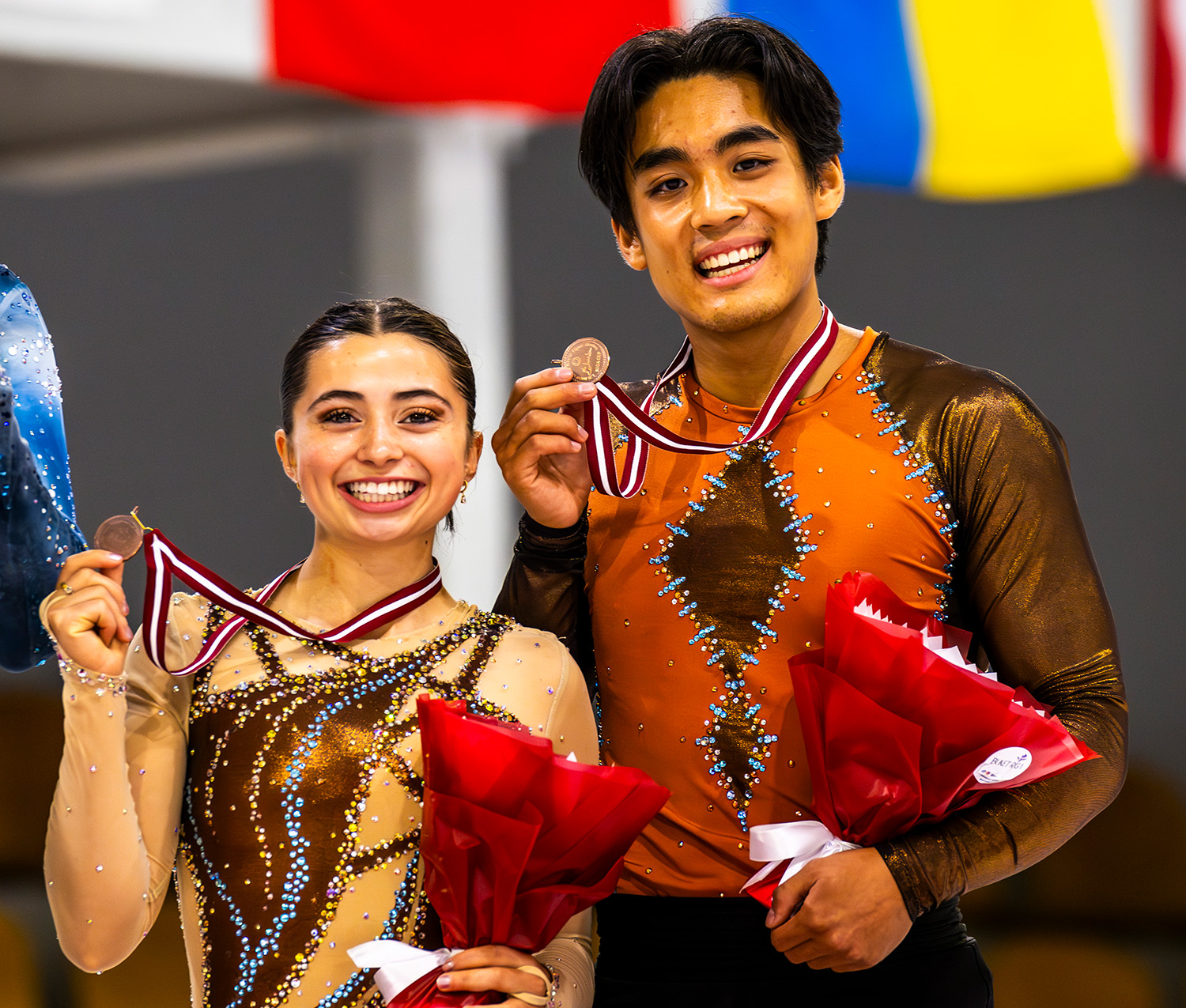 Olivia Flores and Luke Wang hold up their bronze medals with one hand, holding flowers in their other, as they stand on the podium at JGP Latvia. They wear their free skate costumes, which have brown and orange colors.