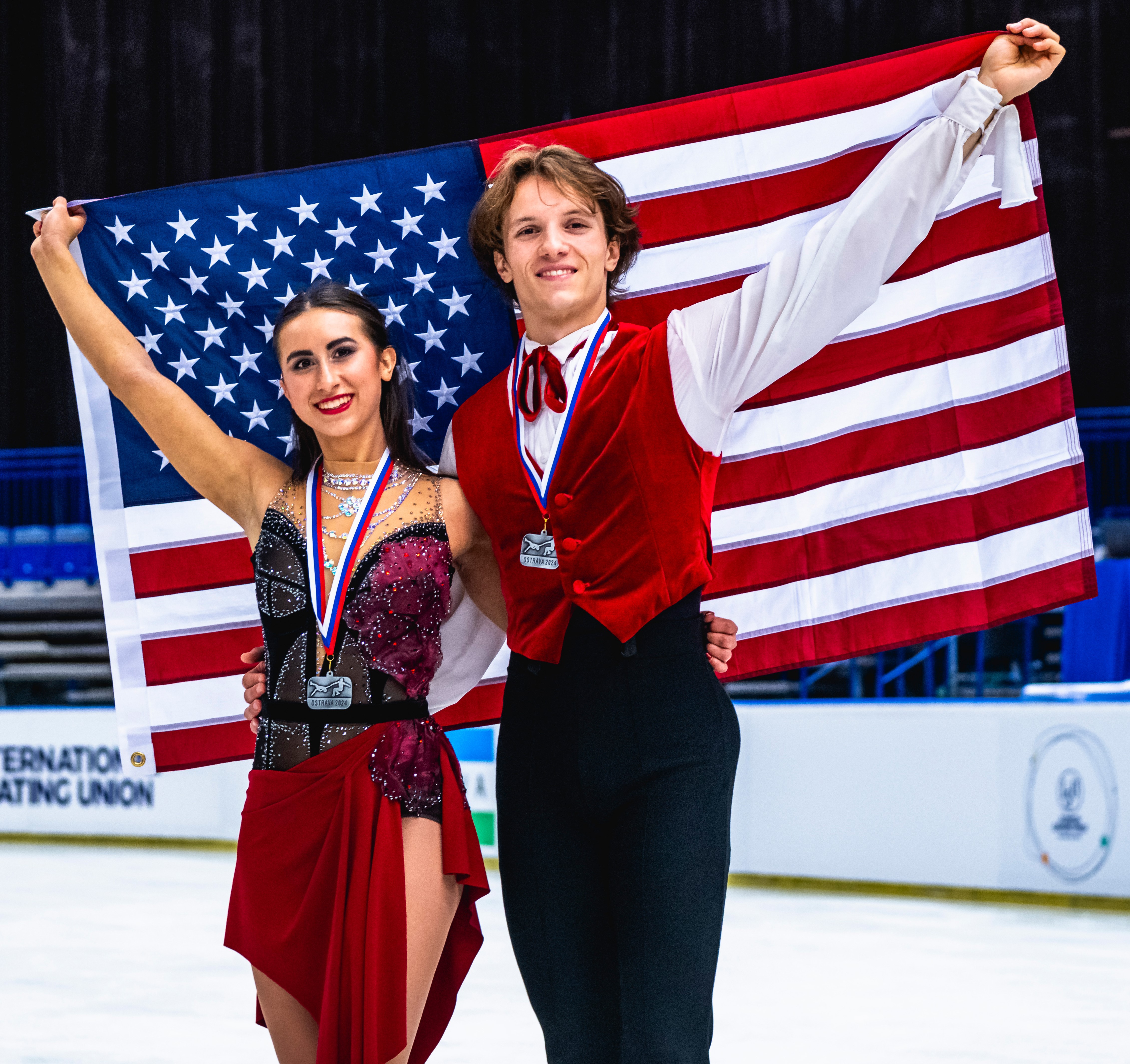 Katarina Wolfkostin and Dimitry Tsarevski pose with their medals and the American flag after taking silver at JGP Czech Republic. Katarina is wearing a red skating dress and Dimitry is wearing a white long sleeve shirt with a red vest and black pants. 