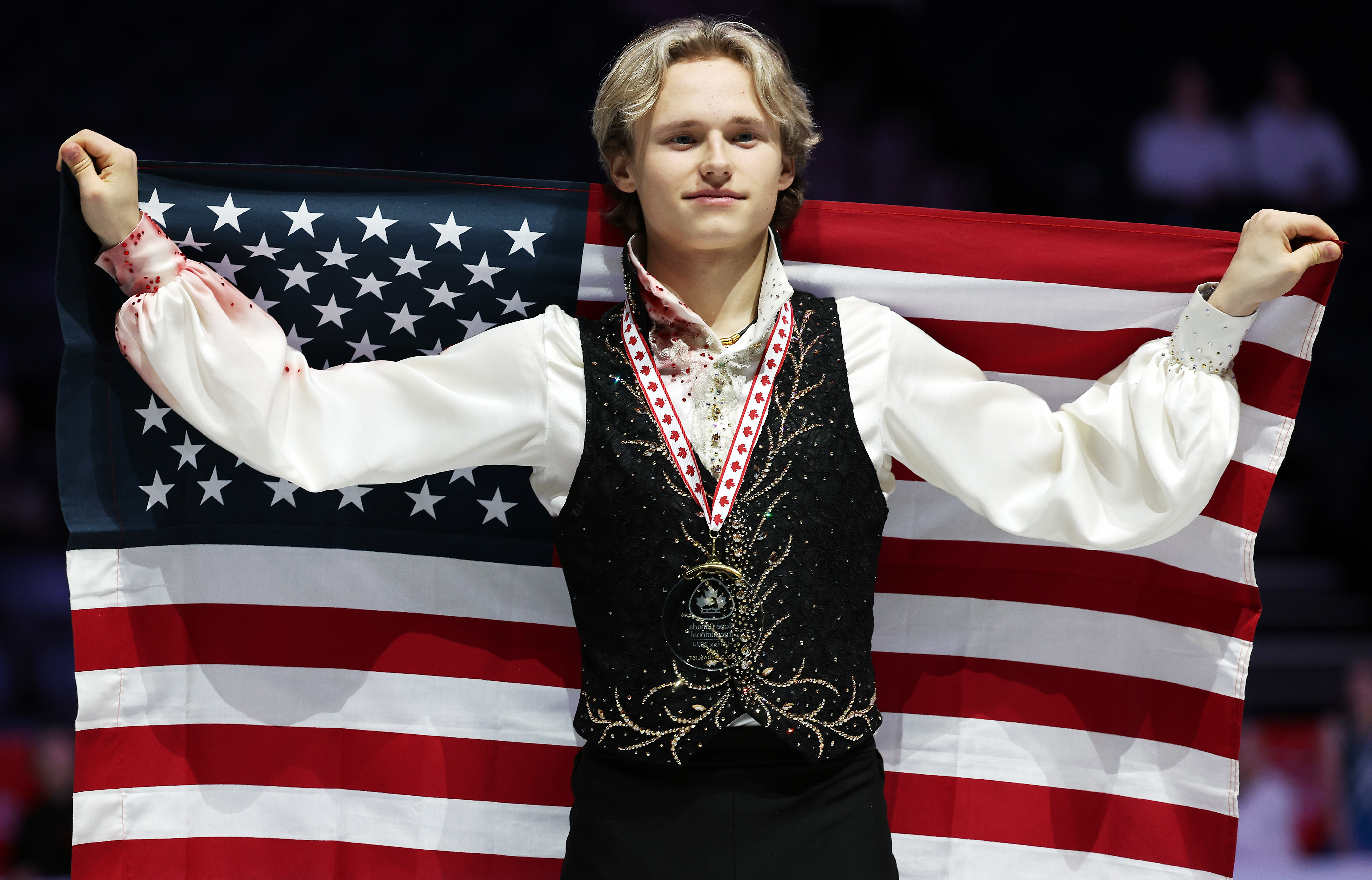 Ilia Malinin, stands on the podium, holding the American flag, at Skate Canada.