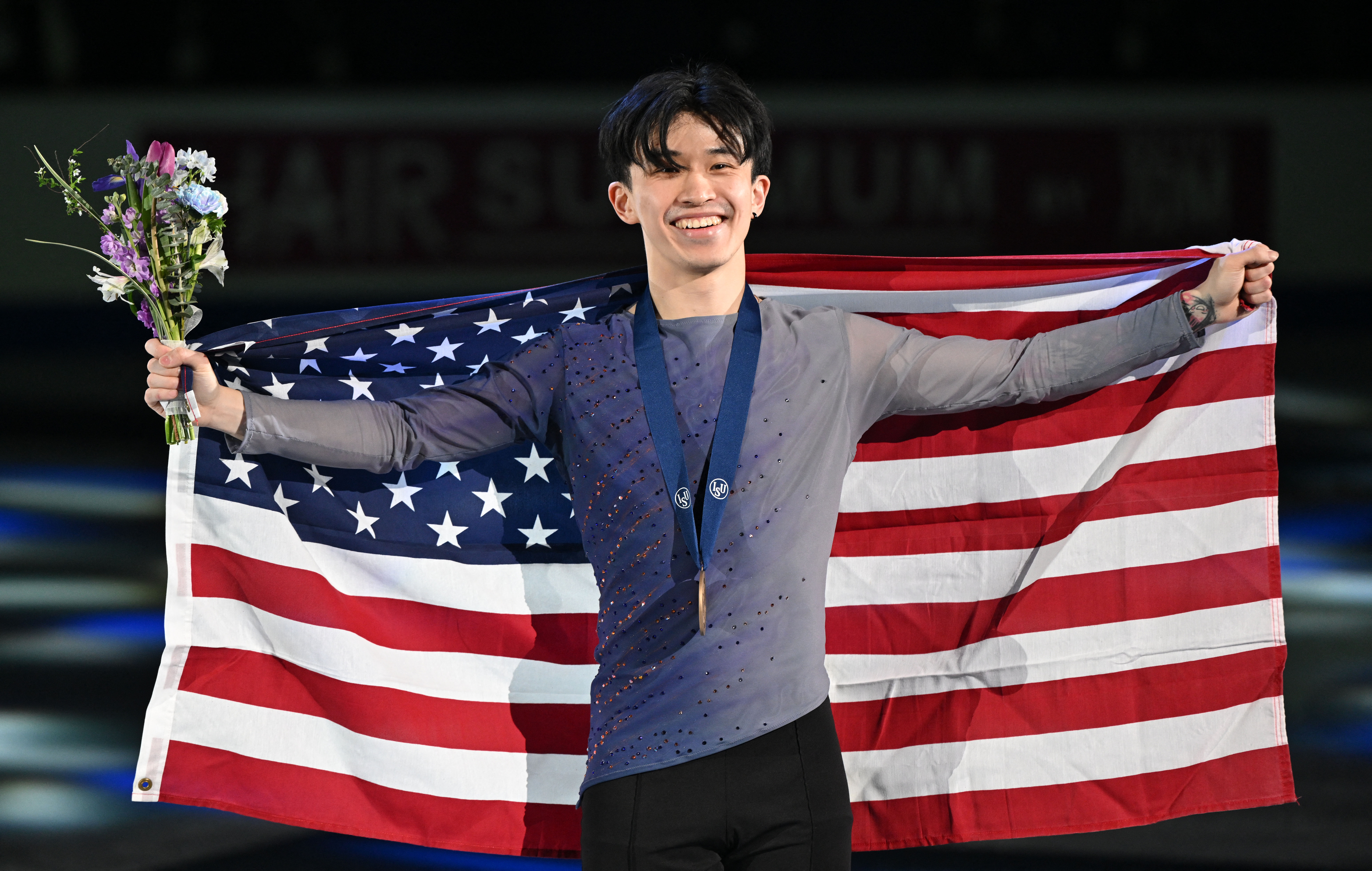 Jimmy Ma poses with his Four Continents bronze medal around his neck while holding an American flag behind him and a bouquet of flowers in his left hand. Jimmy is a young Asian man with short black hair. He is wearing a long sleeve blue and tan ombre top with jewel details on the front and black pants