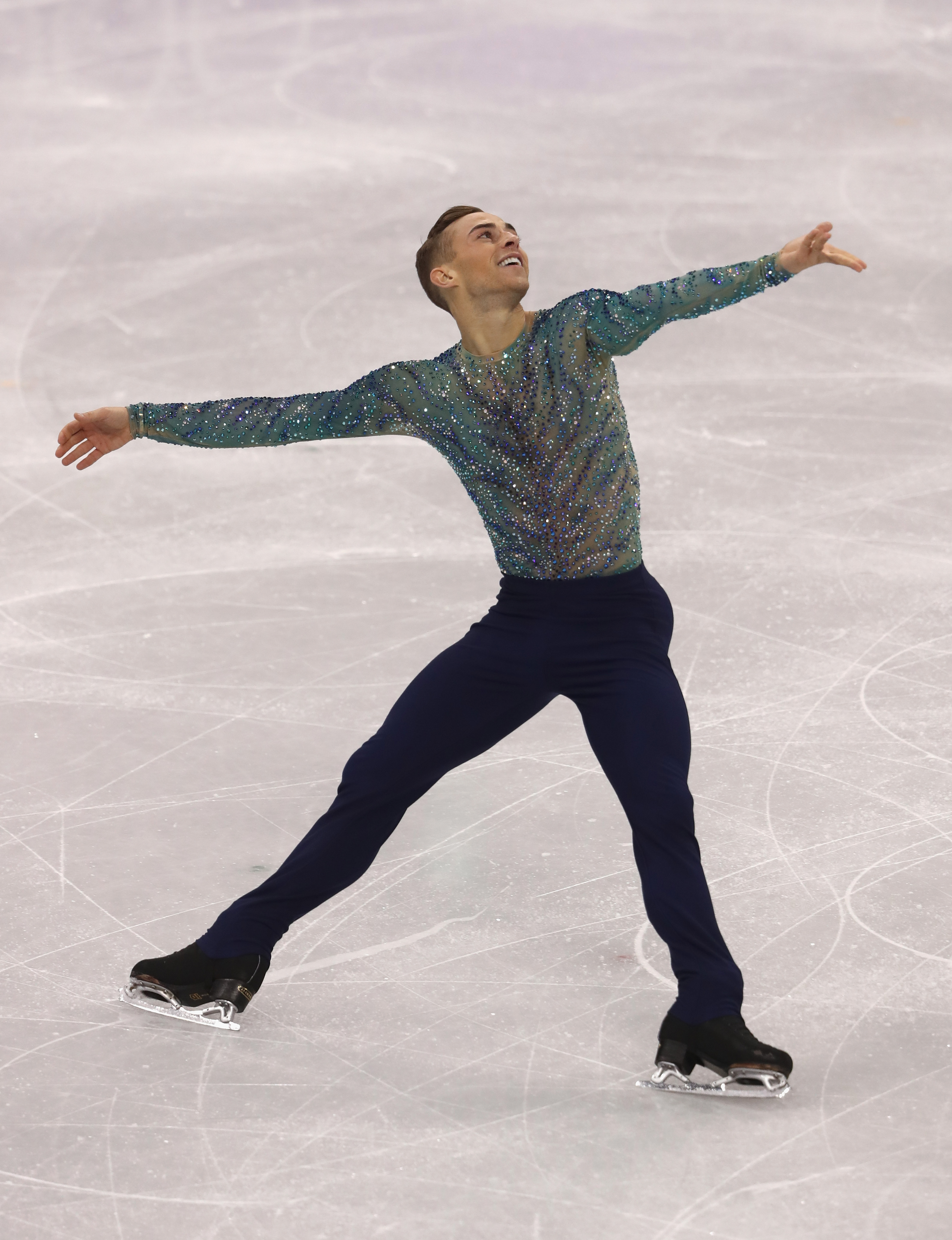 Adam Rippon competes at the 2018 Olympic Winter Games. He looks to the left smiling with his arms outstretched. He is wearing a blue and purple long sleeve top and black pants. 