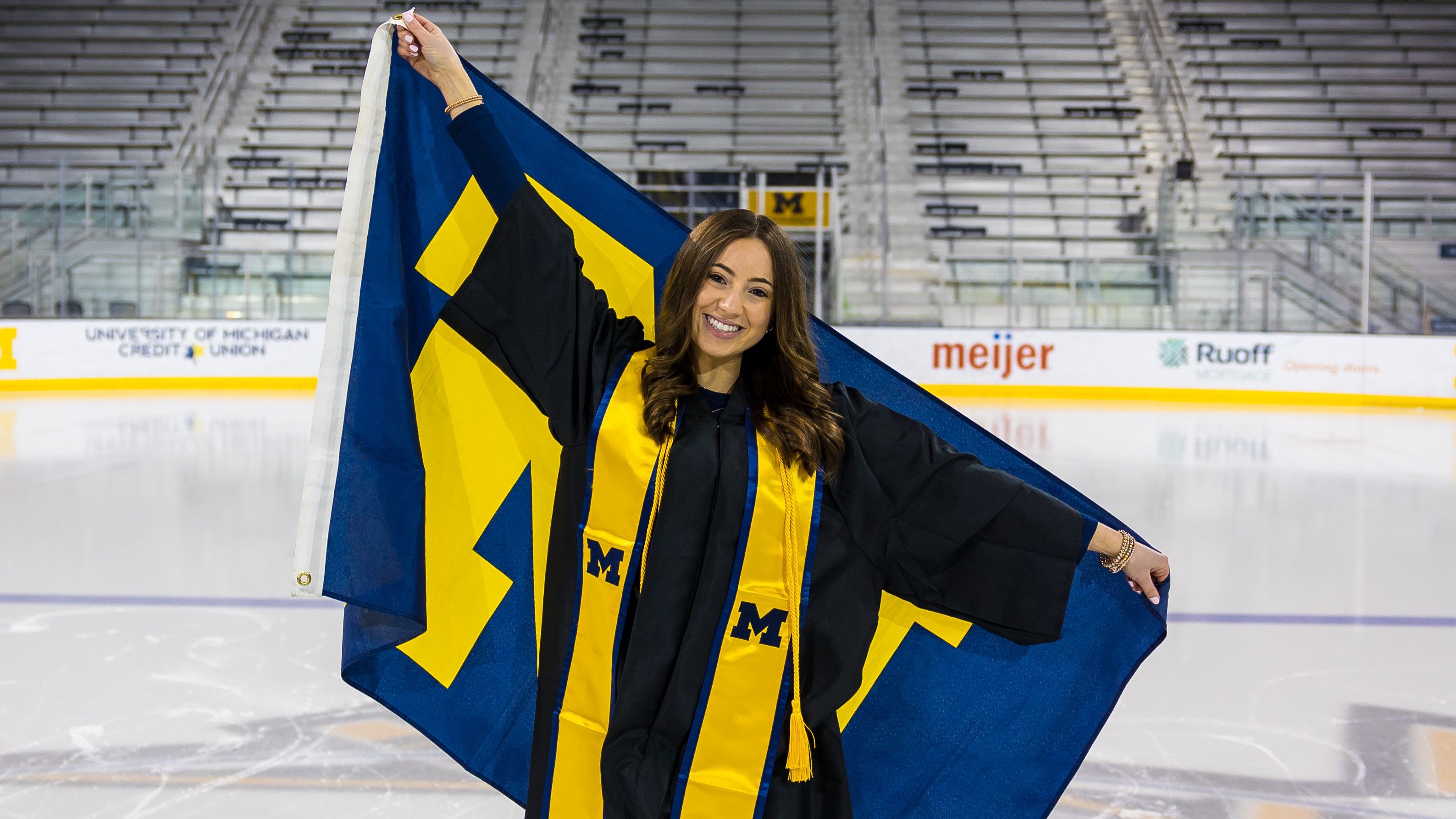 Hailey poses on the ice in her black graduation gown and yellow University of Michigan. Se is holding a blue and yellow University of Michigan flag behind her