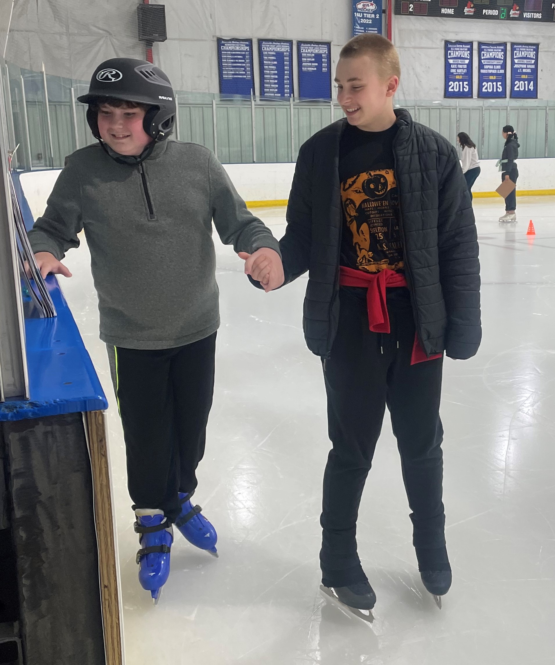 A skater helps a young man with autism balance on the ice. The skater (right) assisting is a teenage boy with short blonde hair wearing a Halloween tshirt, grey jacket and black pants. The boy with autism has on a black helmet, grey quarter zip jacket and black pants. The two hold hands 