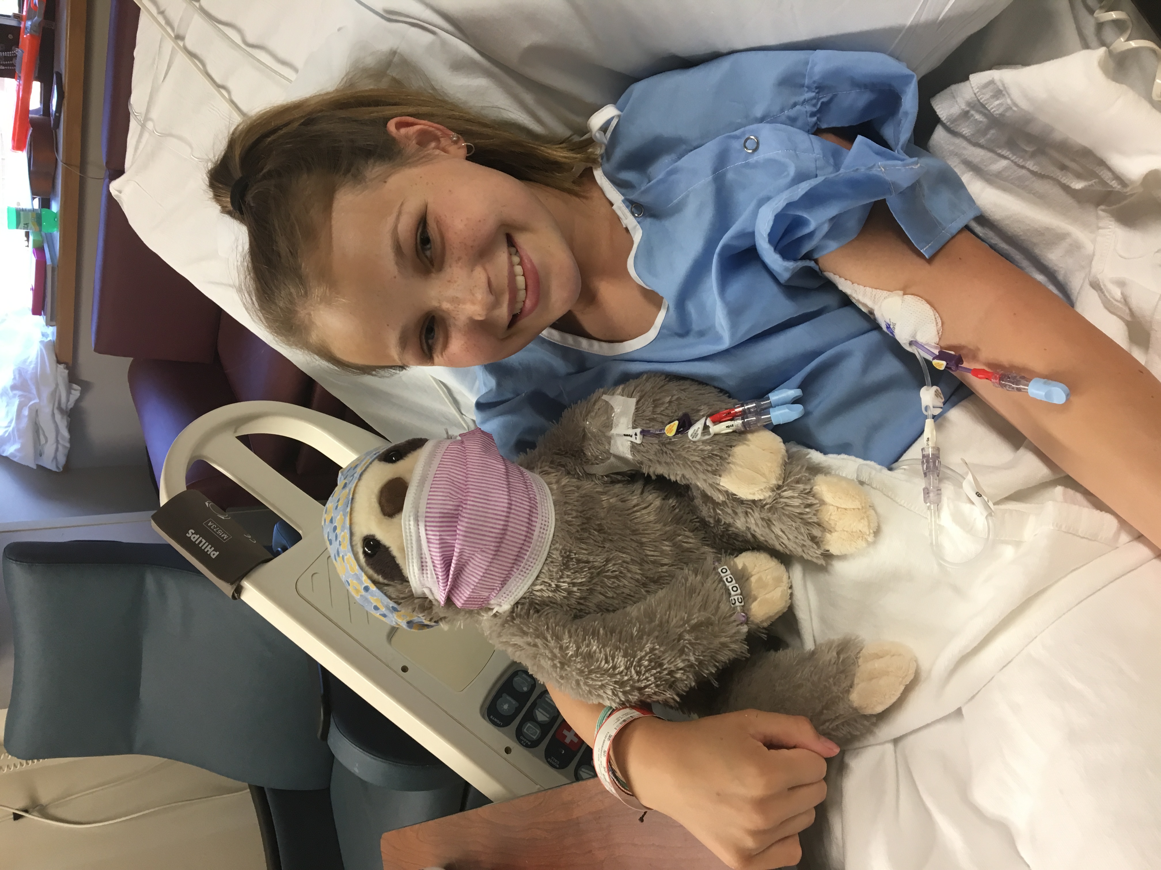 Joelle Hammann smiles from the hospital bed alongside a stuffed sloth wearing a mask.