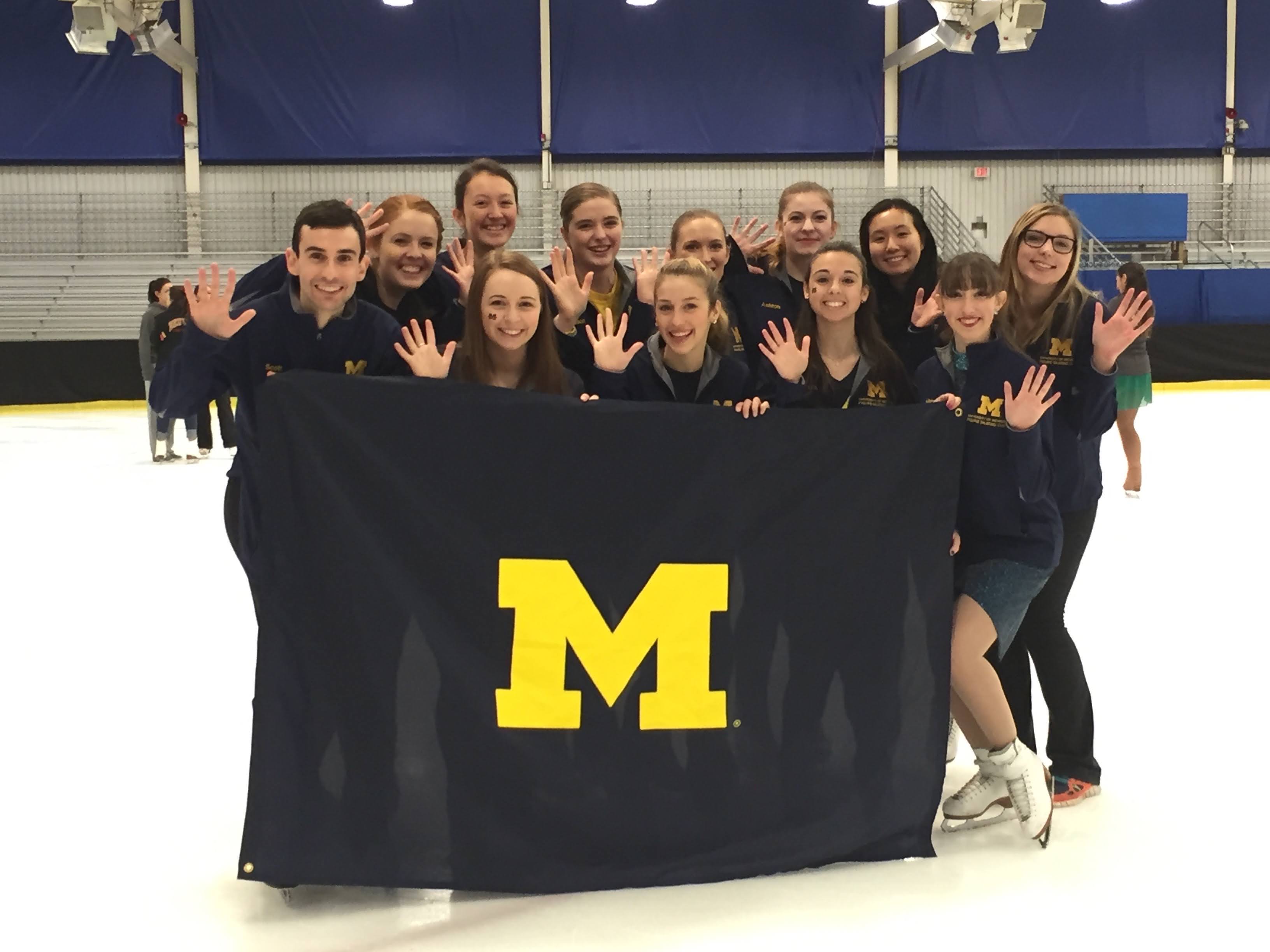 Scott Brody poses with a group of students on the ice. The group is holding a large University of Michigan flag which is blue with a large yellow M in the center. 