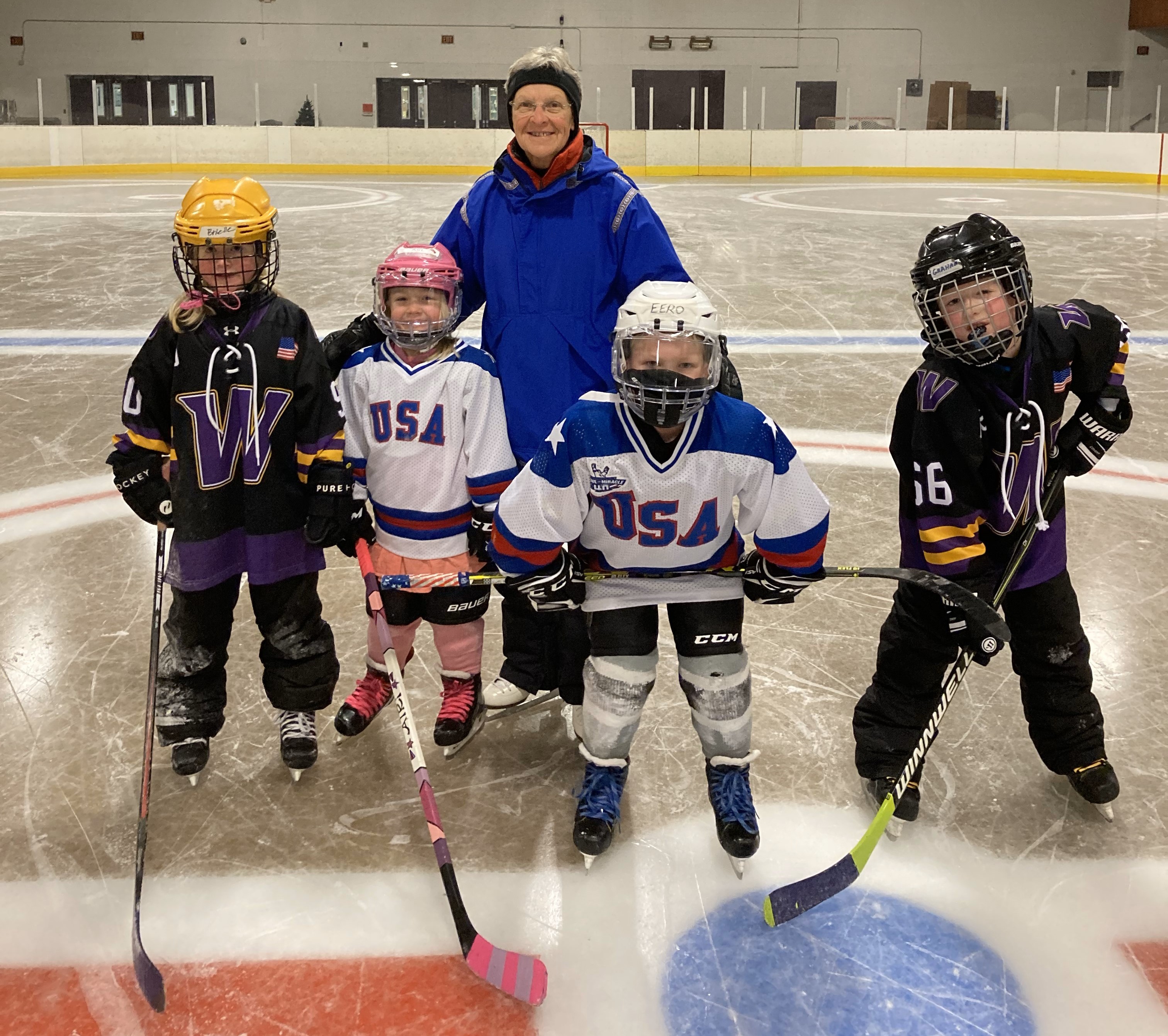 Linda poses for a picture with her four grandkids on the ice. The four grandkids are all wearing hockey gear with two wearing white USA jerseys and the other two wearing black jerseys with a large letter W on the front