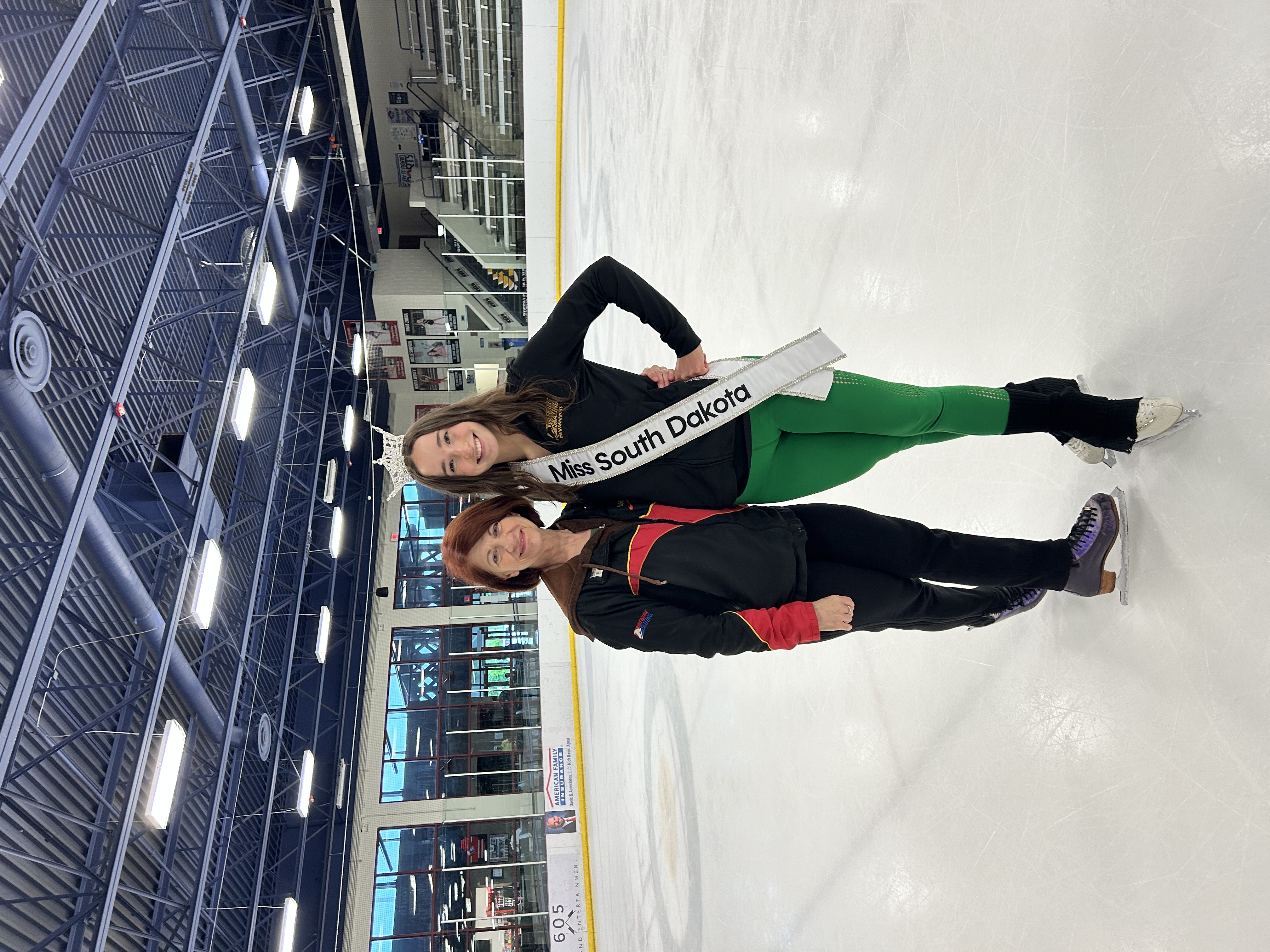 Coach Debbie Minter, wearing black with two red stripes, stands next to Joelle Simpson, dressed in a black top and green pants, with a Miss South Dakota banner over her shoulder.