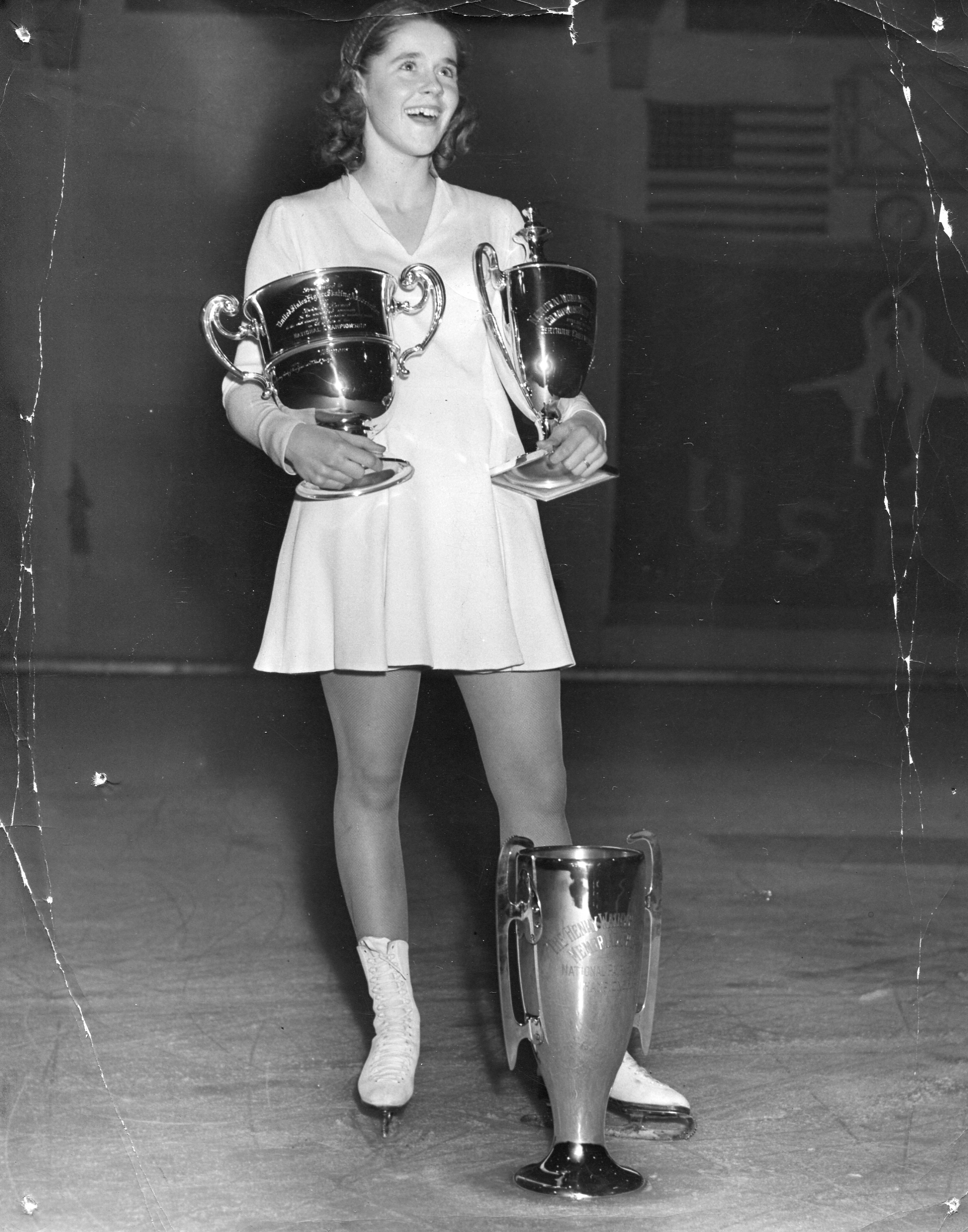 Joan Tozzer stands on the ice in a vintage skating dress holding several trophies.