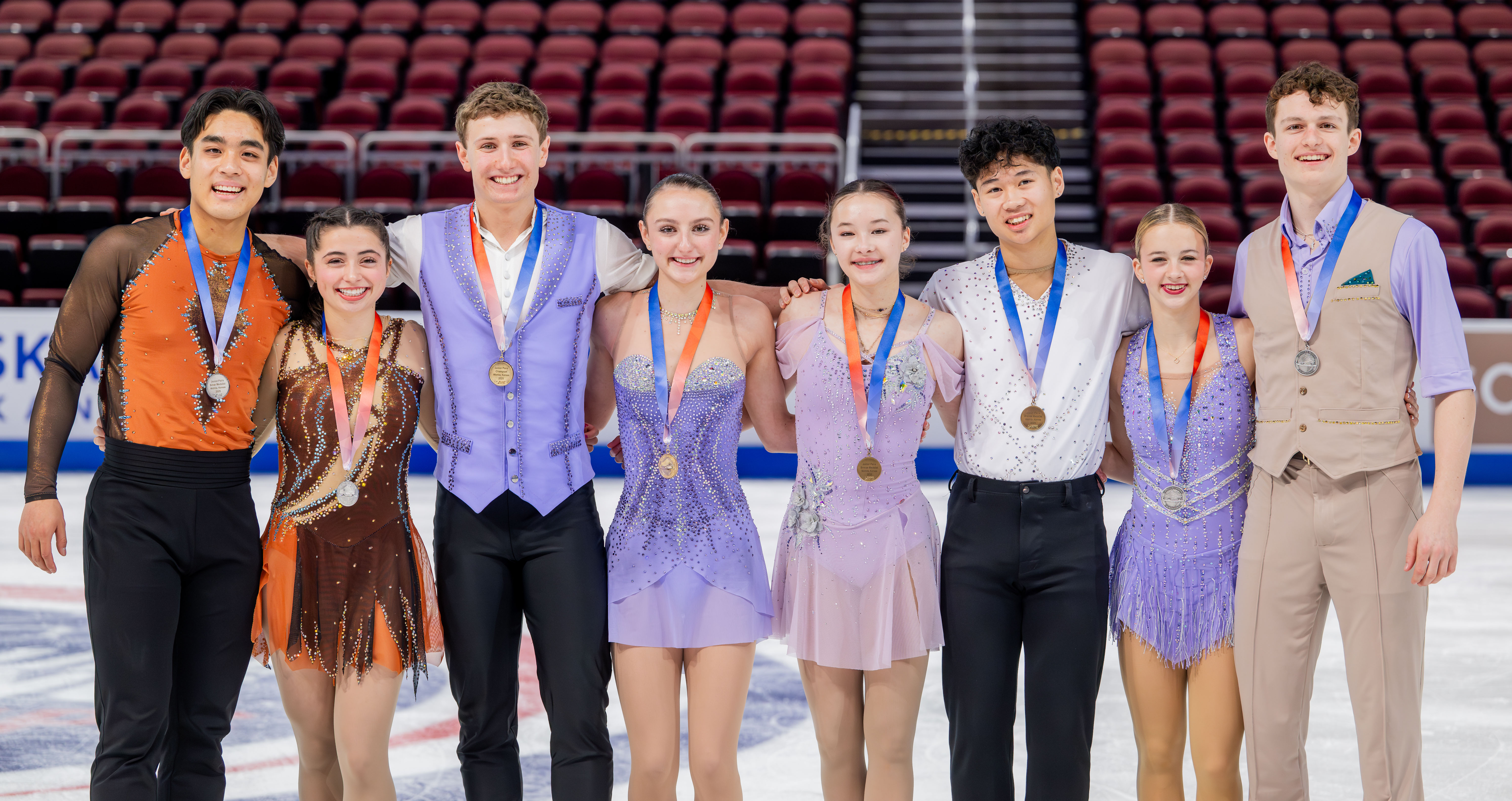 The junior pairs podium stands together on the ice in a line during the victory ceremony, medals around their necks.