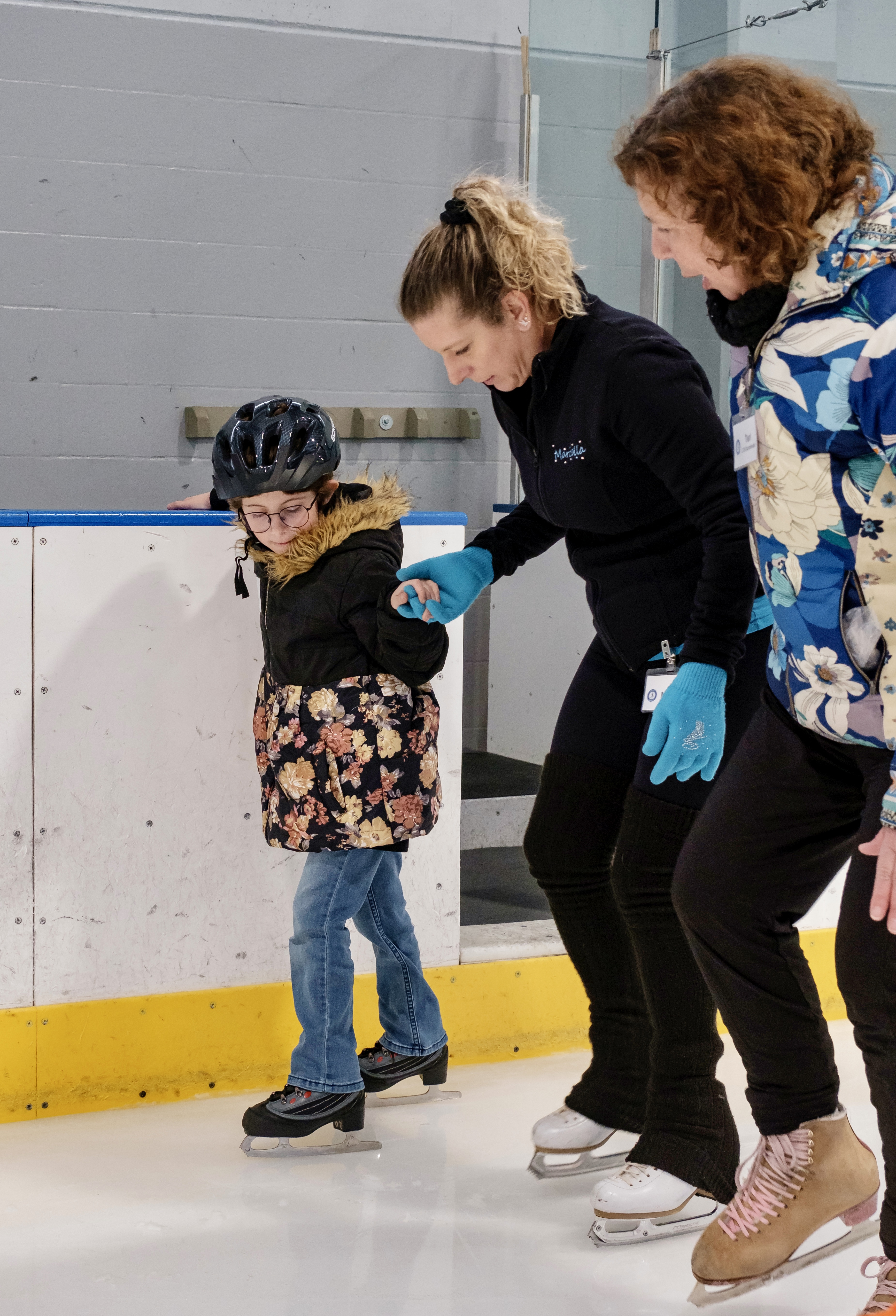 A coach helps a young skater balance on skates. The young skater is a young white girl wearing a black helmet, floral jacket and jeans. The coach standing next to her and holding her hand is an older white woman with blonde hair in a ponytail. She is wearing a black jacket and black pants with blue gloves. Standing to the other side of the coach is another adult woman. She has shoulder length red hair and is wearing a blue floral jacket with black pants