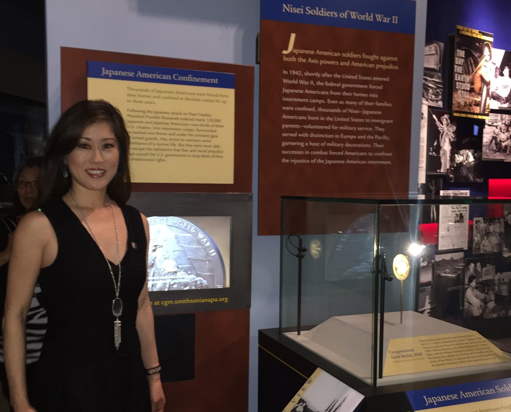 Kristi poses in front of the Smithsonian’s Congressional Gold Medal Exhibit in Washington, D.C.. Kristi is an Asian woman with long black hair wearing a black cocktail dress 