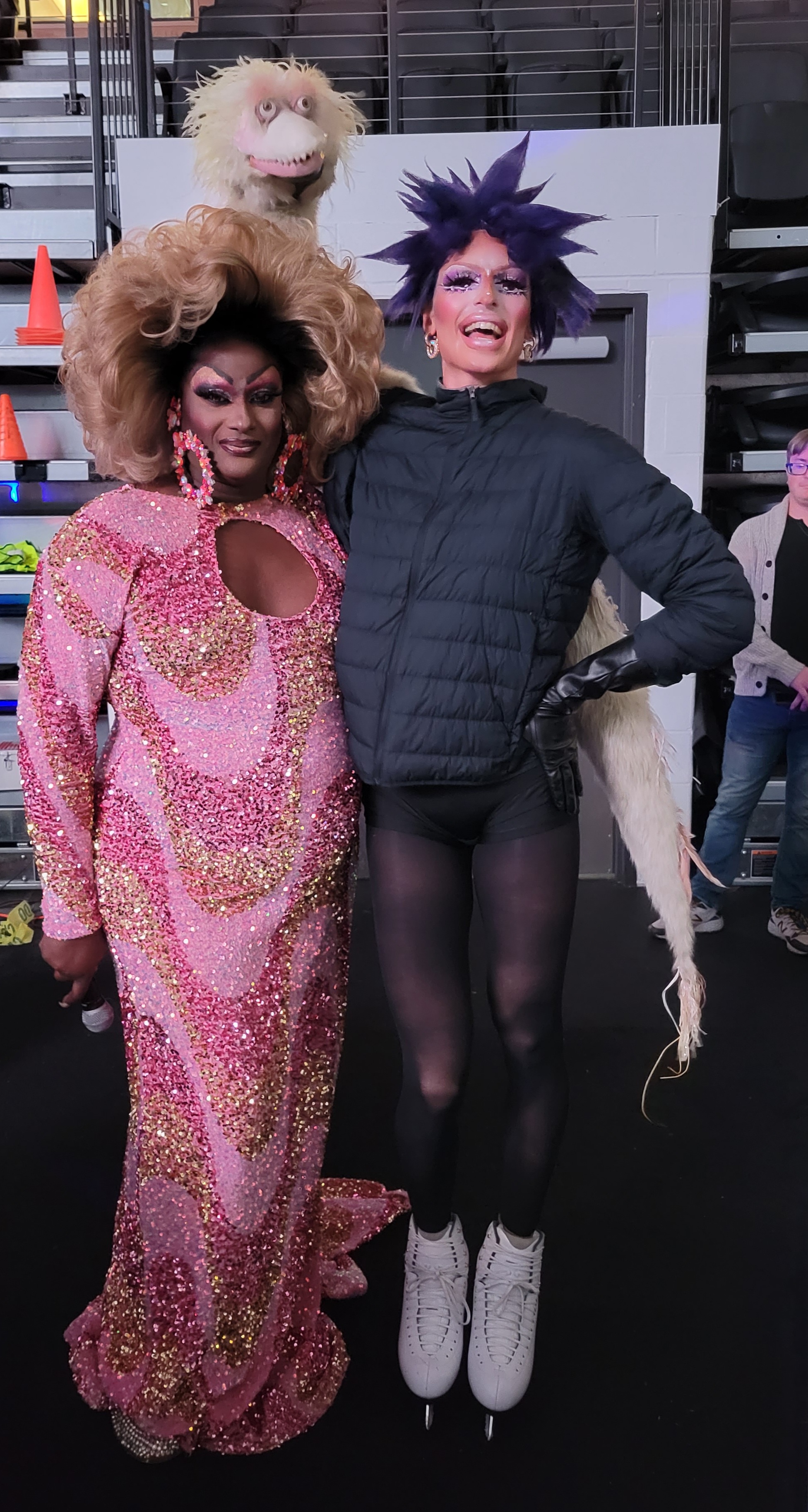 Drag queens Mizery McRae (left) and (milk) pose for a photo. McRae is a black woman wearing a long sleeved and bedazzled pink gown and a large blonde afro wig. Milk is wearing a wig with short and spiky purple hair, a winter jacket, tights and skates