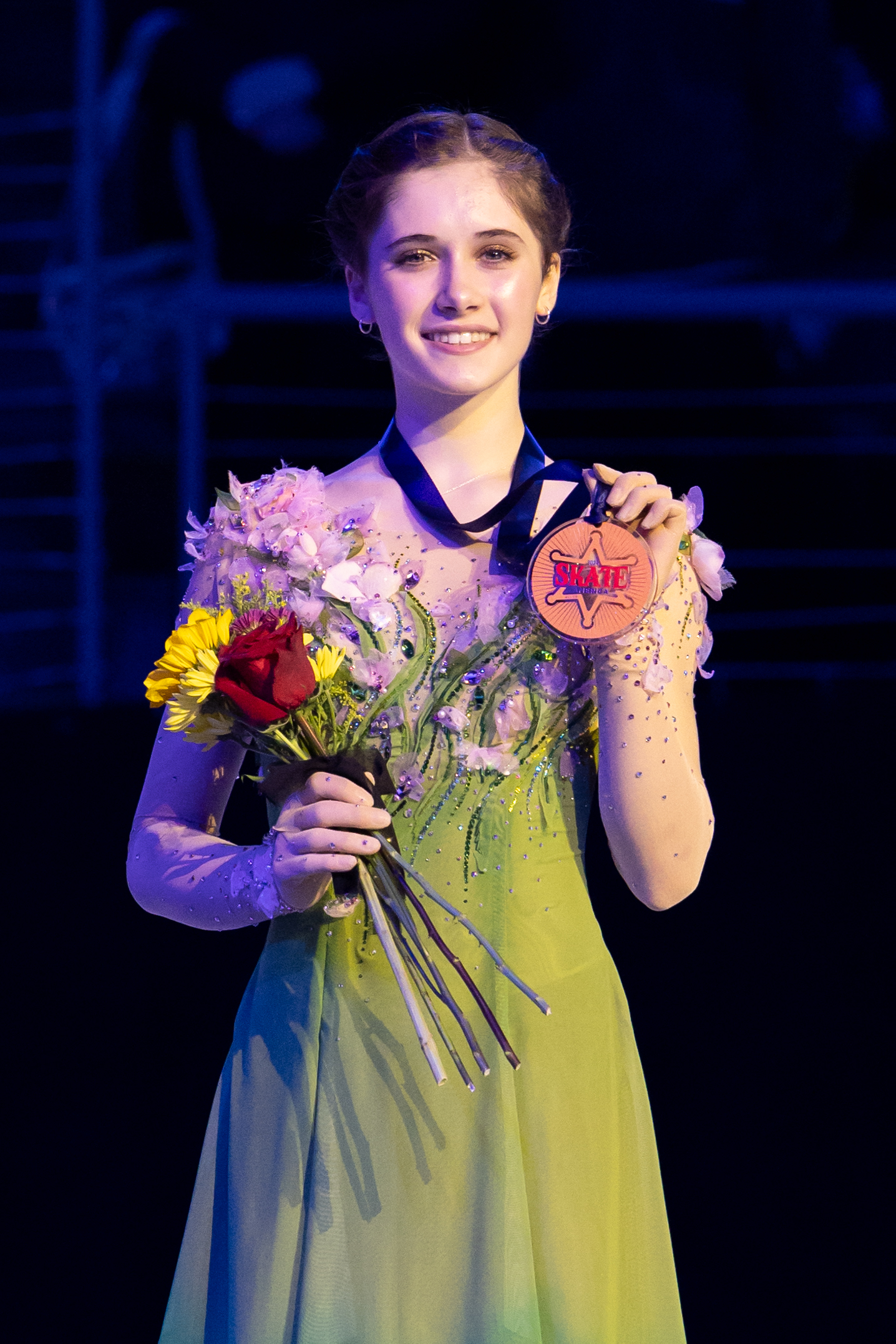 Isabeau Levito, in a green dress, holds up her Skate America medal while holding flowers and smiles while standing on the podium at 2024 Skate America.