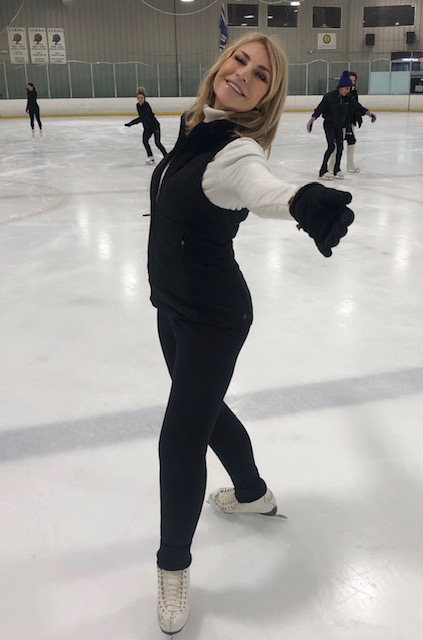 Photo of Lorraine on the ice. She is a Caucasian woman with shoulder length blonde hair. She is wearing a white long sleeve shirt with a black vest on top and black pants. She is standing with her arms outstretched to her sides