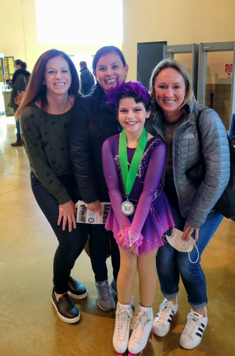 Hannah smiles while urrounded by some of her nurses from when she was in treatment at her first competition since starting treatment. Hannah is a young girl wearing a purple skating costume with a matching headband. She has a medal with a green ribbon around her neck