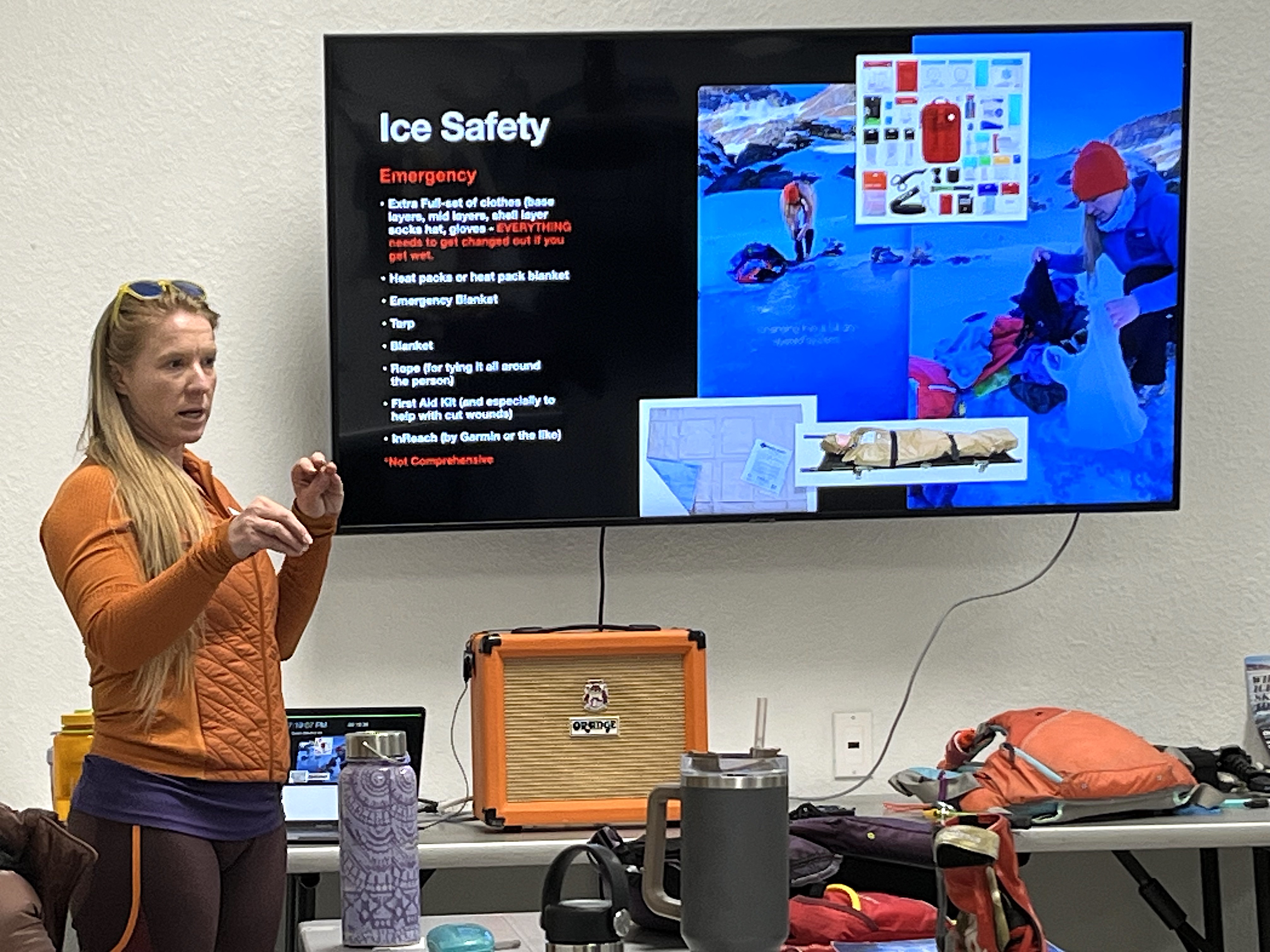 Laura stands in front of a tv talking to her students with  Powerpoint slide titled Ice Safety on the screen. She has long blonde hair and is wearing a love sleeve orange shirt