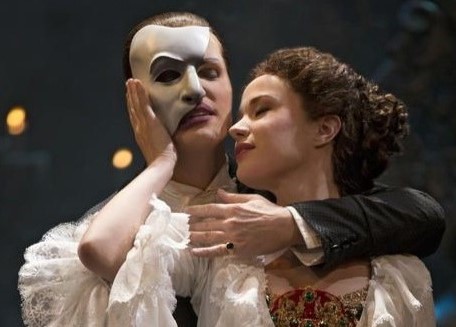 Sierra Boggess, in a white, ruffled dress, embraces the Phantom who has a white mask covering half of his face.