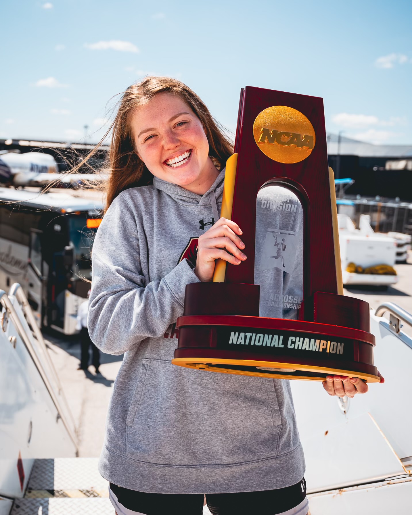 Maddy poses with an NCAA championship trophy. The trophy is wood with glass in the front. She has long brown hair and she is wearing a grey sweatshirt