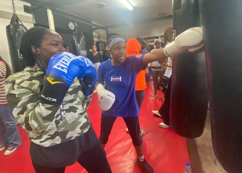 A member of Figure Skating in Harlem is taught how to fit a punching bag from a member of the organization Fight With Insight. She FSH athlete is wearing a camo jacket and the teacher has on a blue tshirt and black pants